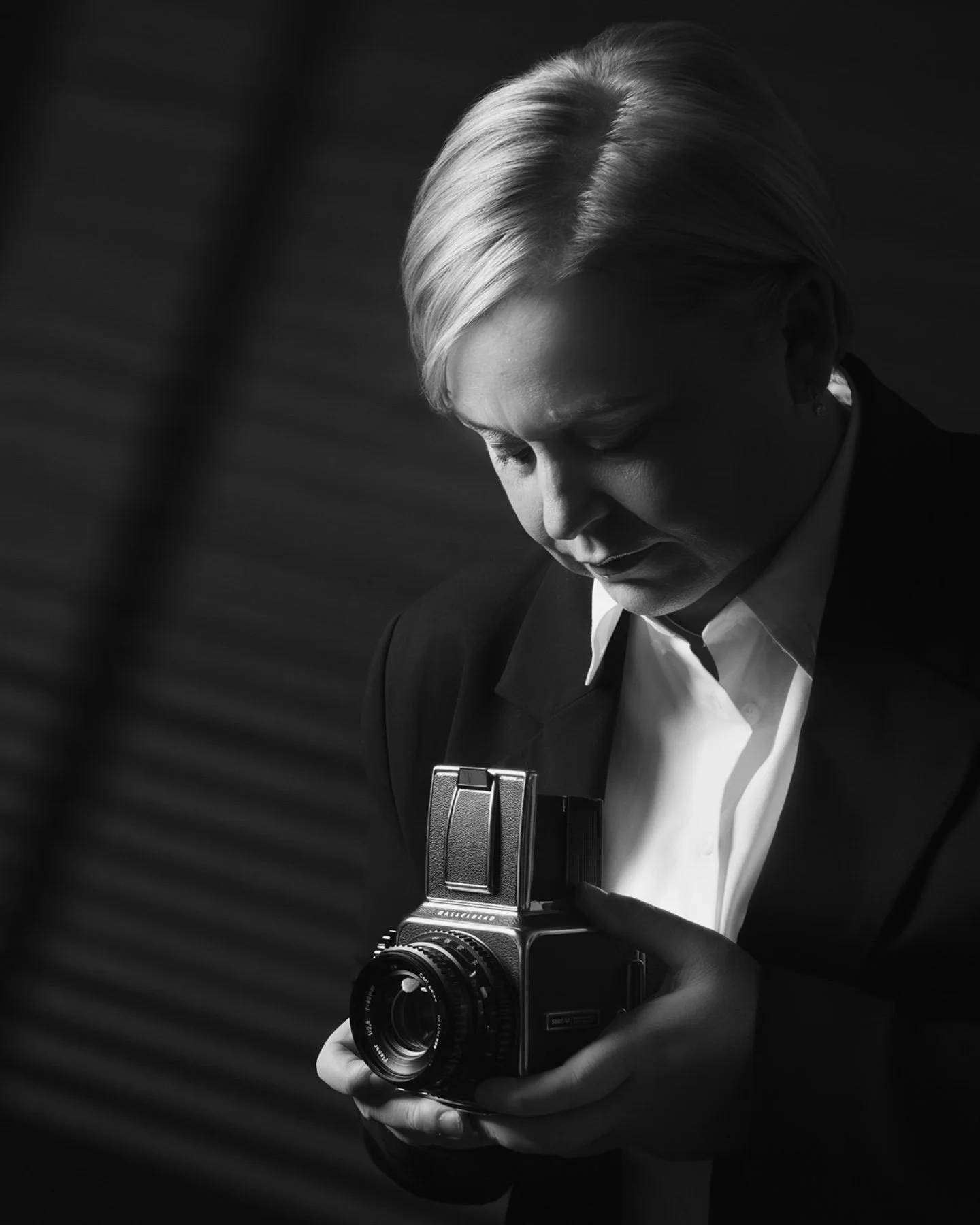 A black-and-white photo of a woman with short blonde hair, dressed in a suit, looking down at a vintage camera she is holding with both hands.