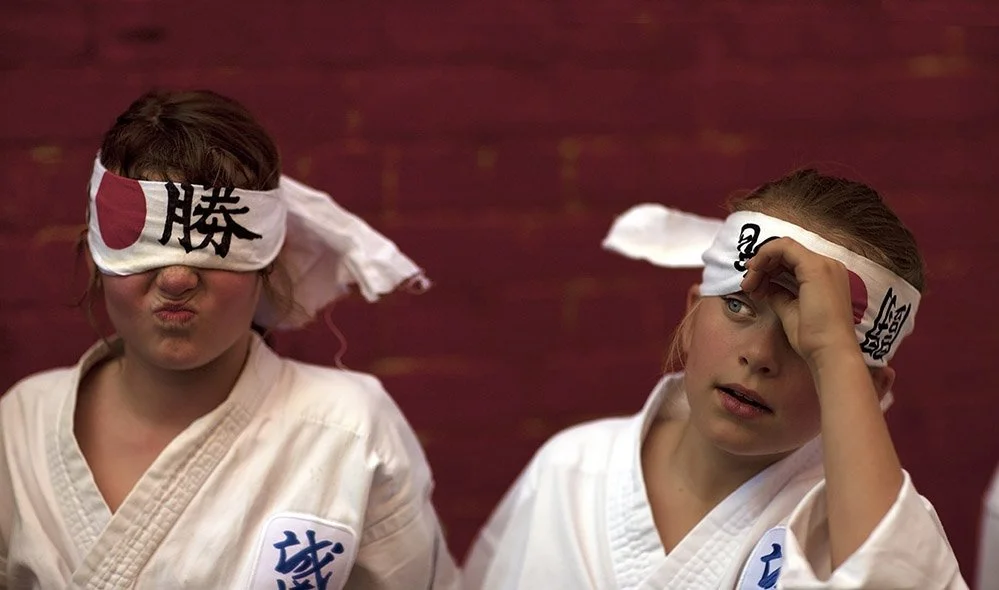 Two young girls in martial arts uniforms wearing headbands with Japanese characters, one with her eyes covered and the other shielding her eyes, sitting against a brick wall.