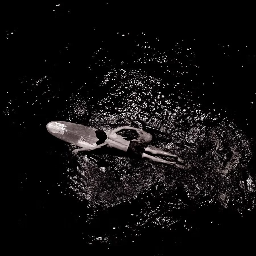 A person lying on a surfboard on dark water, possibly at night, creating ripples.