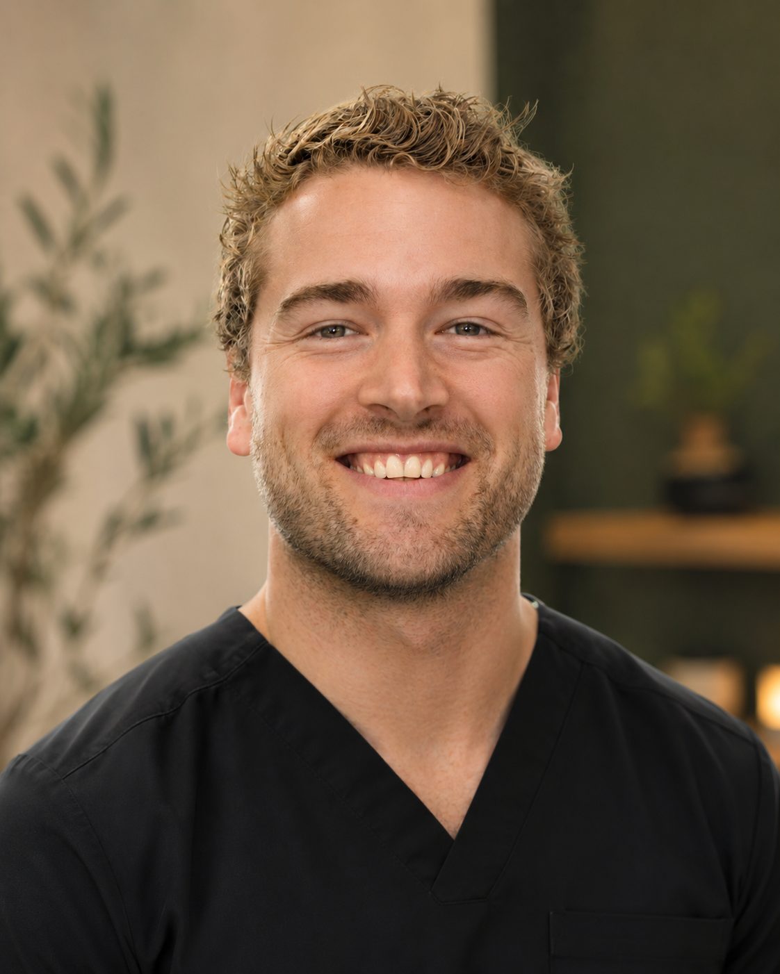 Smiling man with short, curly blonde hair and a beard, wearing black scrubs, in a warmly lit room with plants and decor in the background.