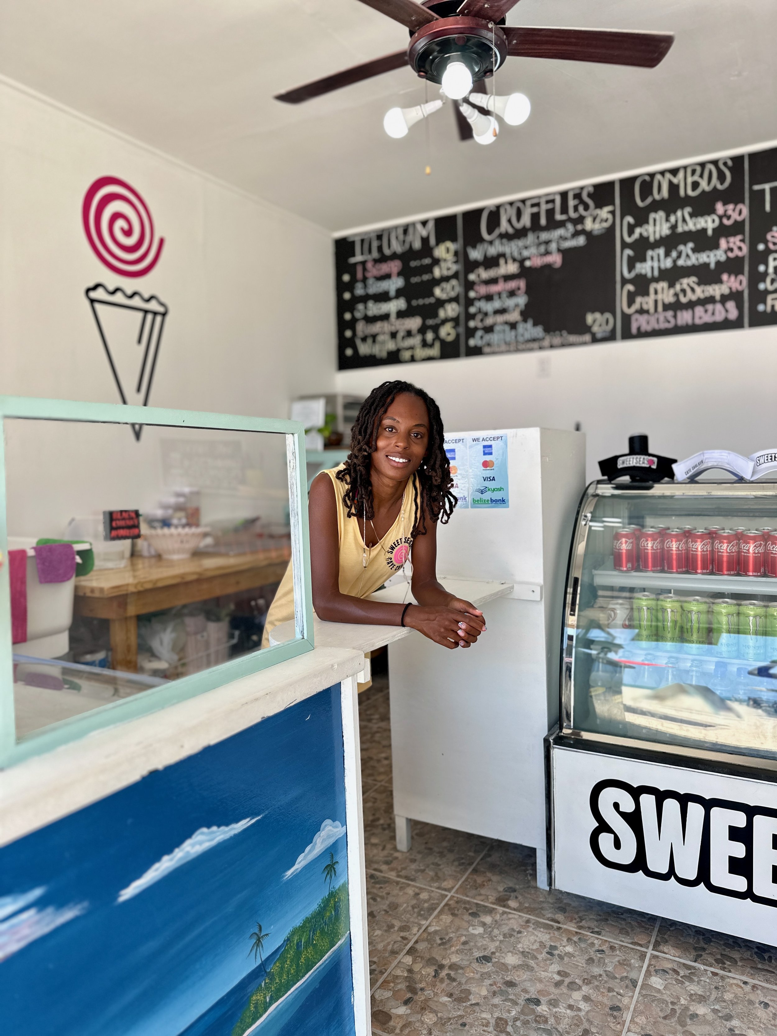 A woman standing behind an ice cream and beverage counter in a shop. The shop has a blackboard menu with items and prices, a ceiling fan with lights, and painted wall art of a pink swirl and an ice cream cone. The woman has dreadlocks and is smiling at the camera.