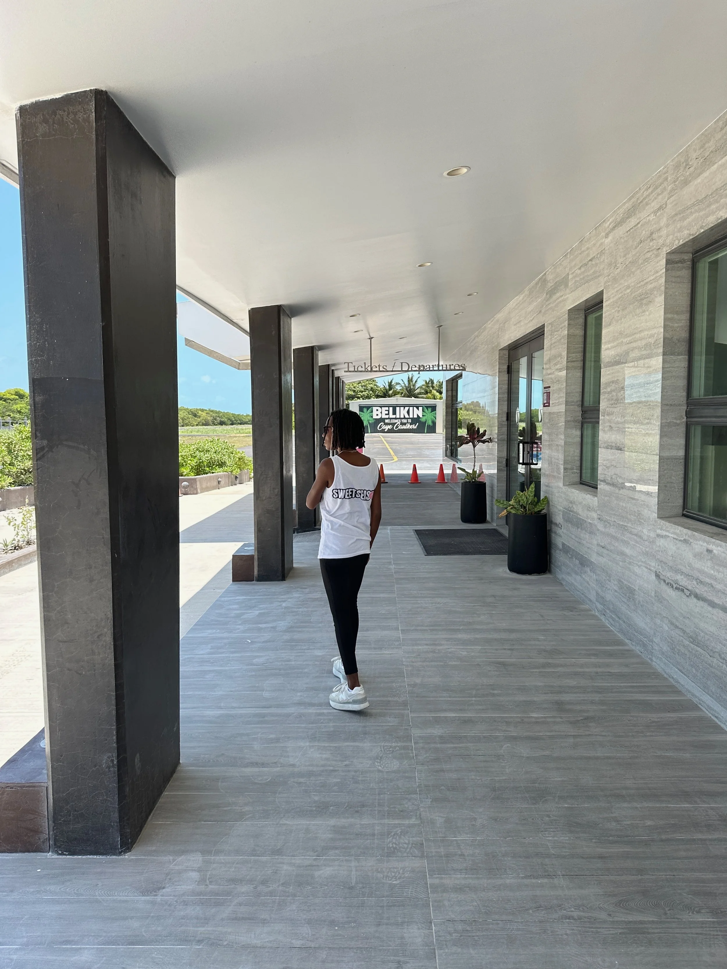 A woman with short black hair, wearing glasses, a white tank top with 'SWEETSHO' written on it, black pants, and white sneakers, stands under a modern building's overhang with black pillars and large windows, facing away from the camera.