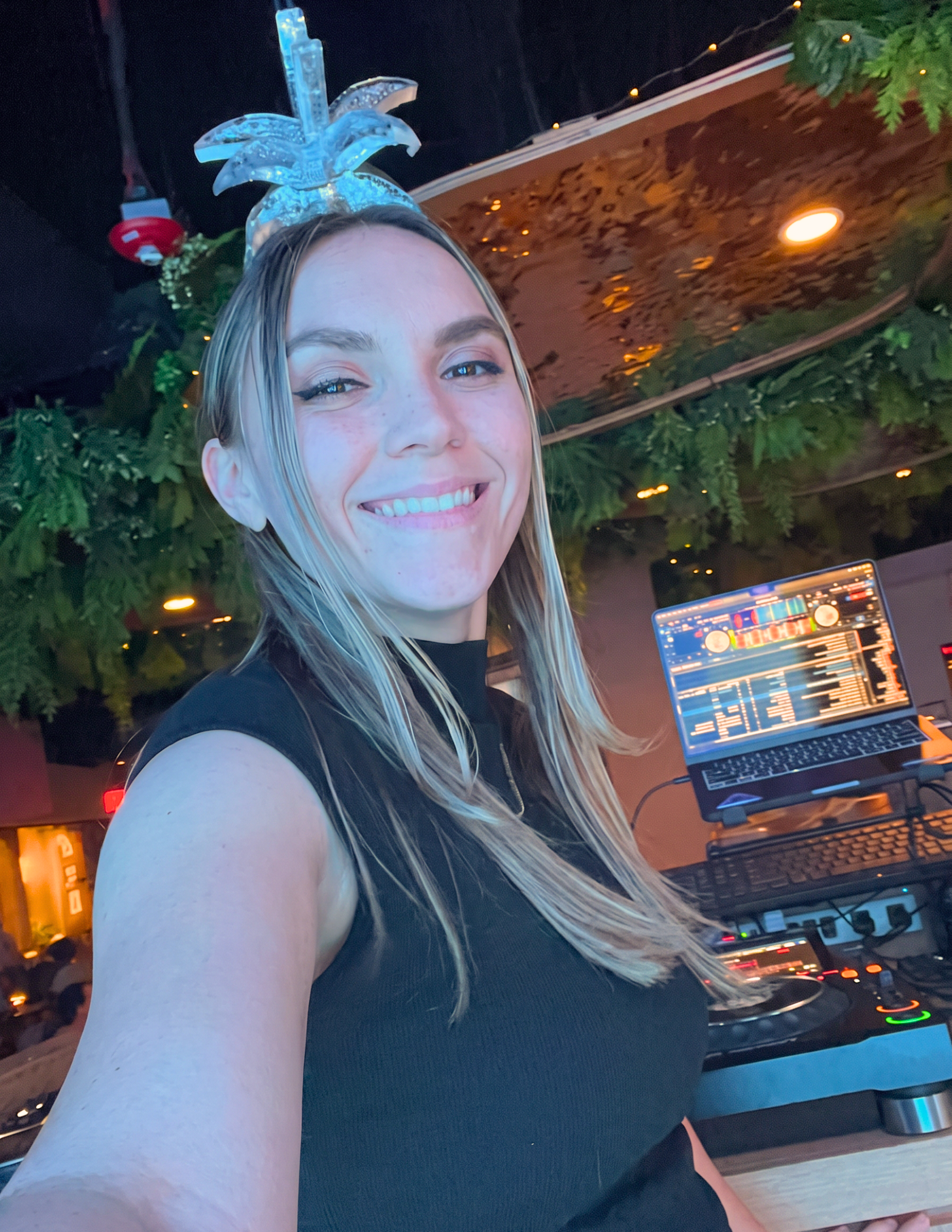 A woman with blonde hair smiling at a DJ setup in a dimly lit venue decorated with greenery and fairy lights.