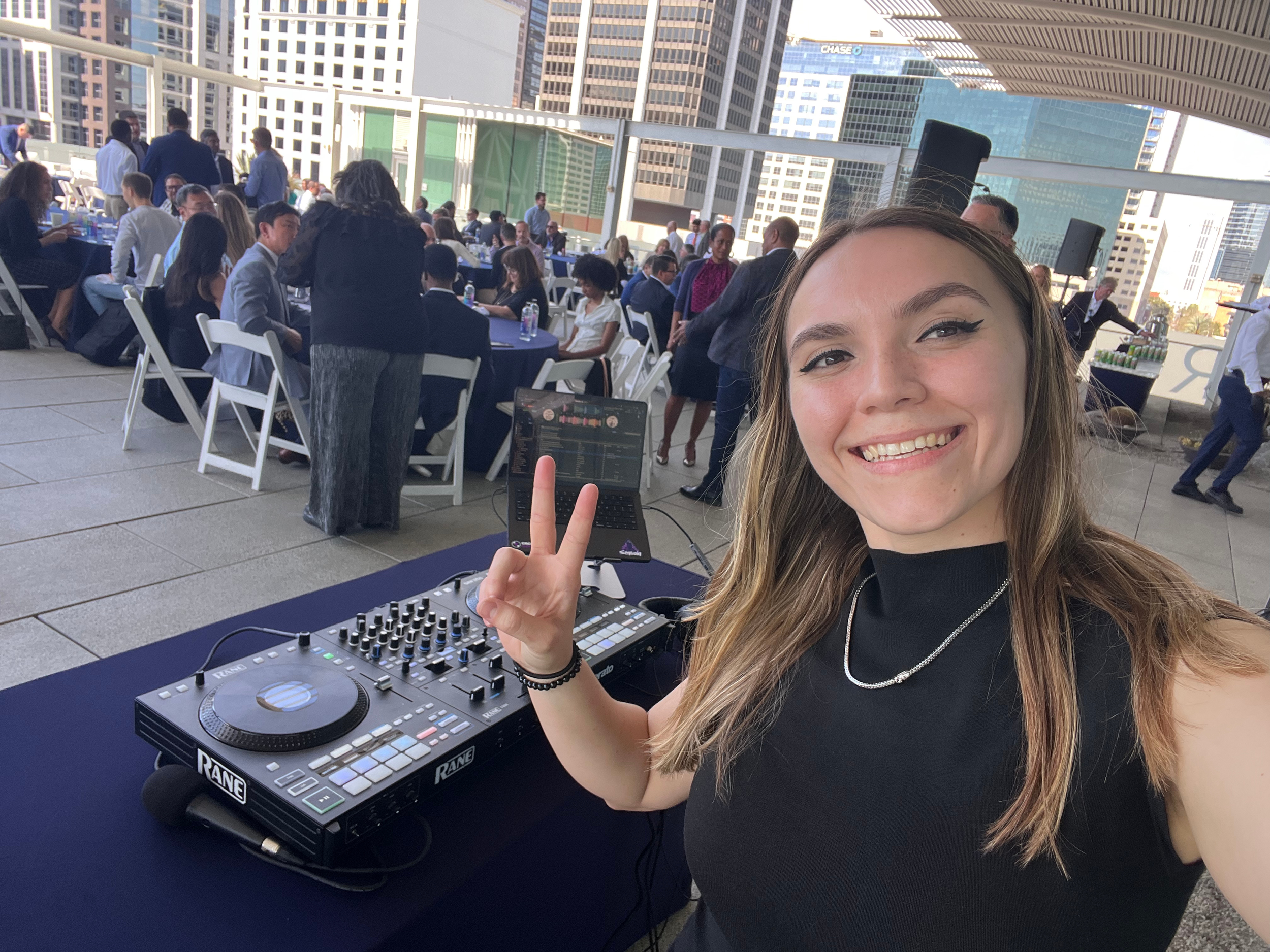 A young woman with long hair smiling at the camera, making a peace sign with her hand, with DJ equipment on a table in front of her at an outdoor rooftop event in a city with tall buildings in the background.