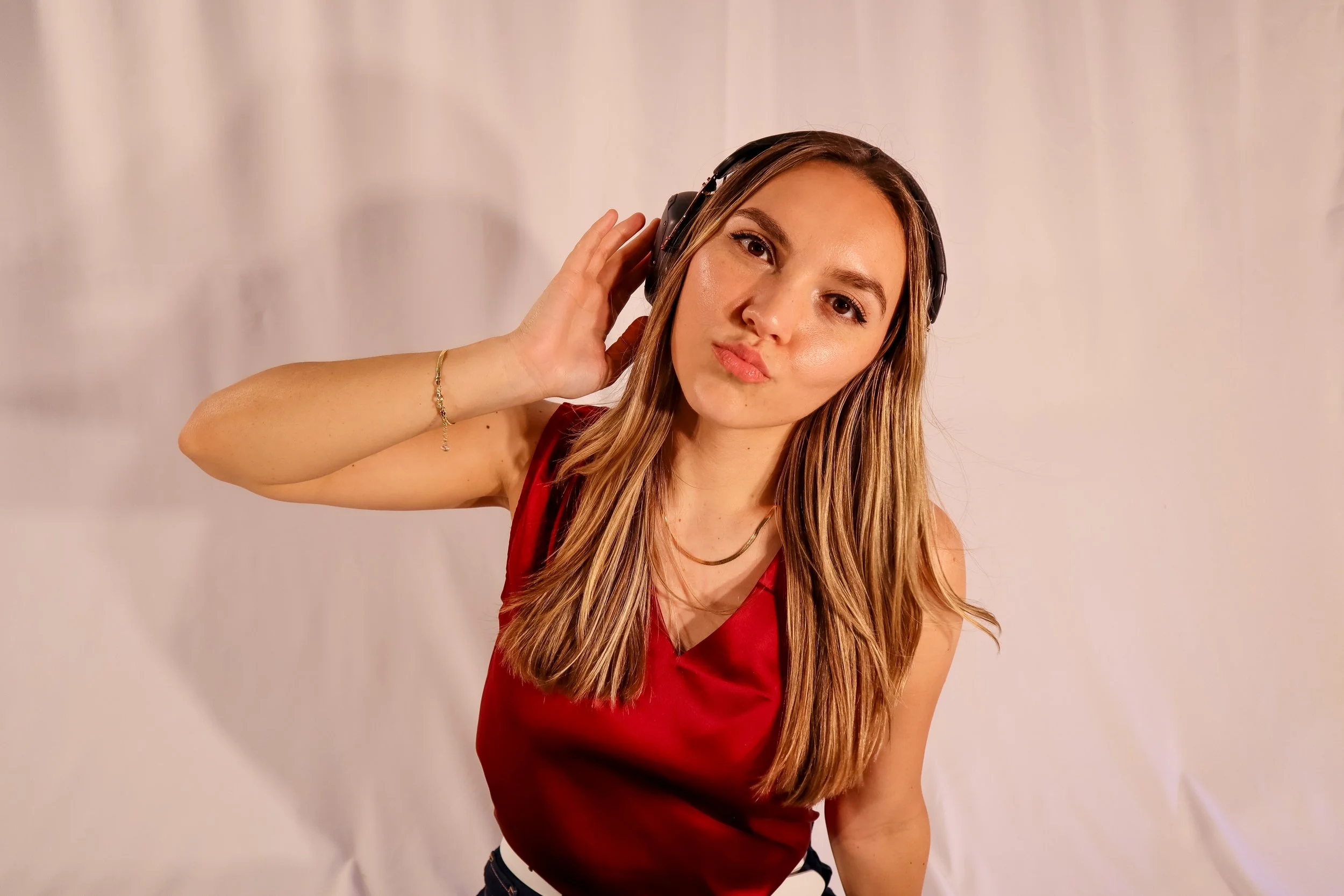A young woman with long, light brown hair is wearing a red sleeveless top and headphones, posing with a slight pout and raising one hand near her ear against a plain light-colored backdrop.