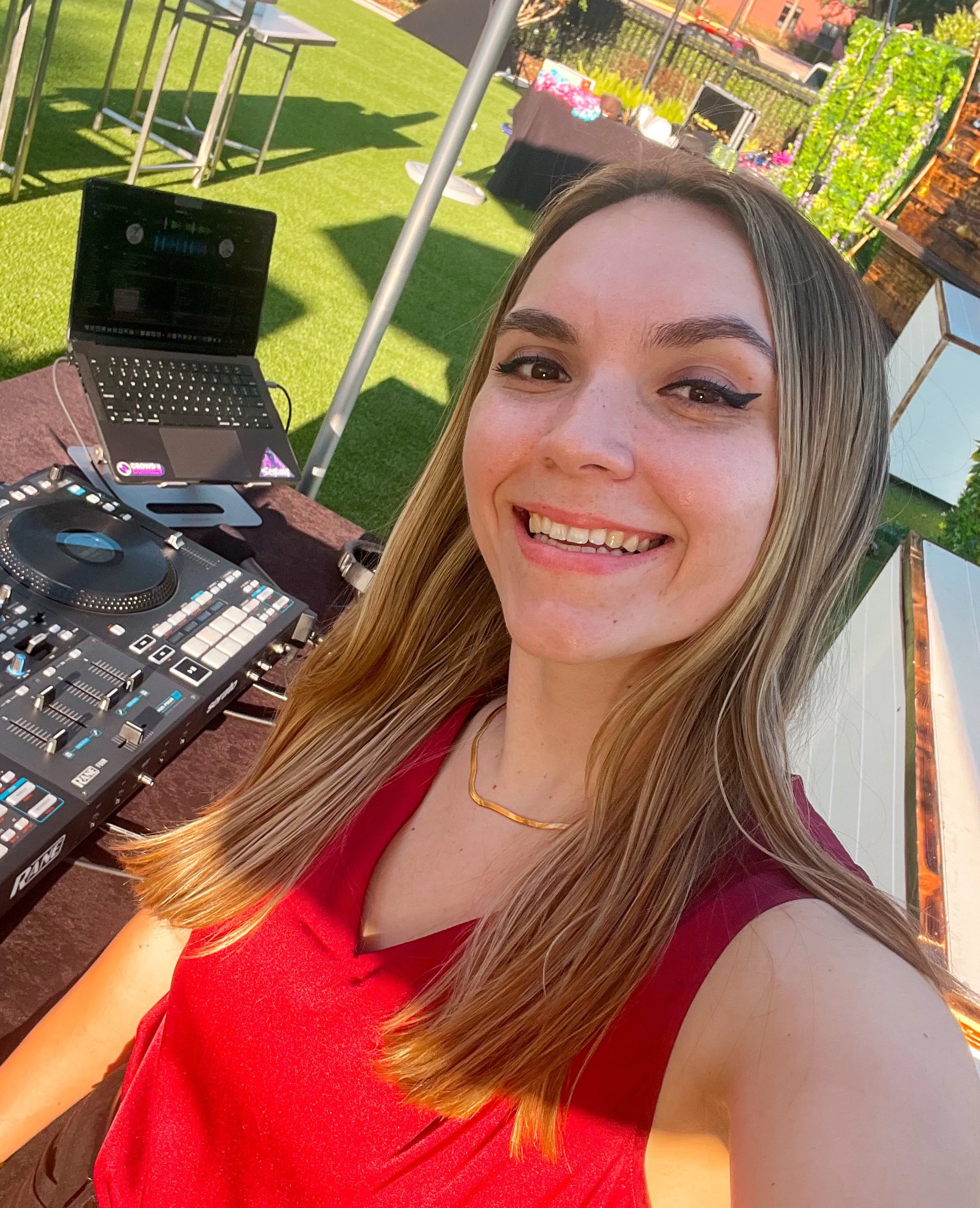 A woman smiling outdoors at a bar or DJ setup with DJ equipment, a laptop, and a table with drinks. There is a grassy area with chairs and tables, and some trees and buildings in the background.