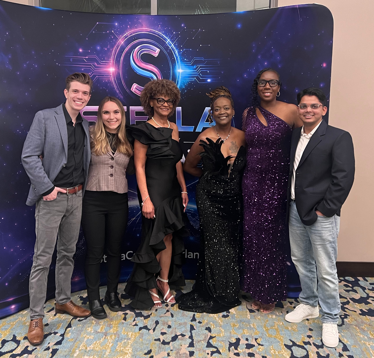 Group of seven people standing together in front of a Starla event backdrop, dressed elegantly for a formal occasion.