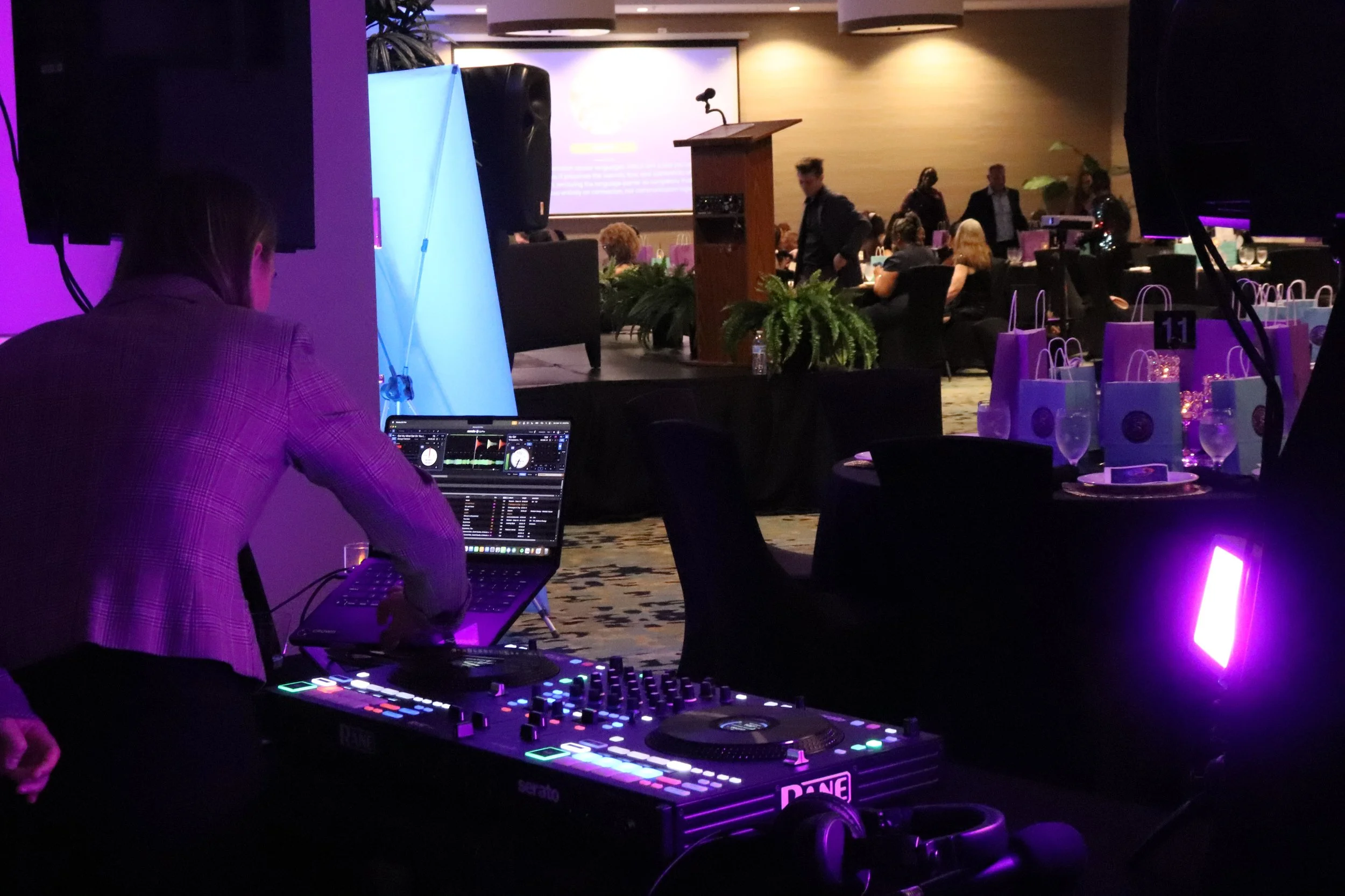 DJ performing with a laptop and DJ equipment at an event with tables and people in the background.