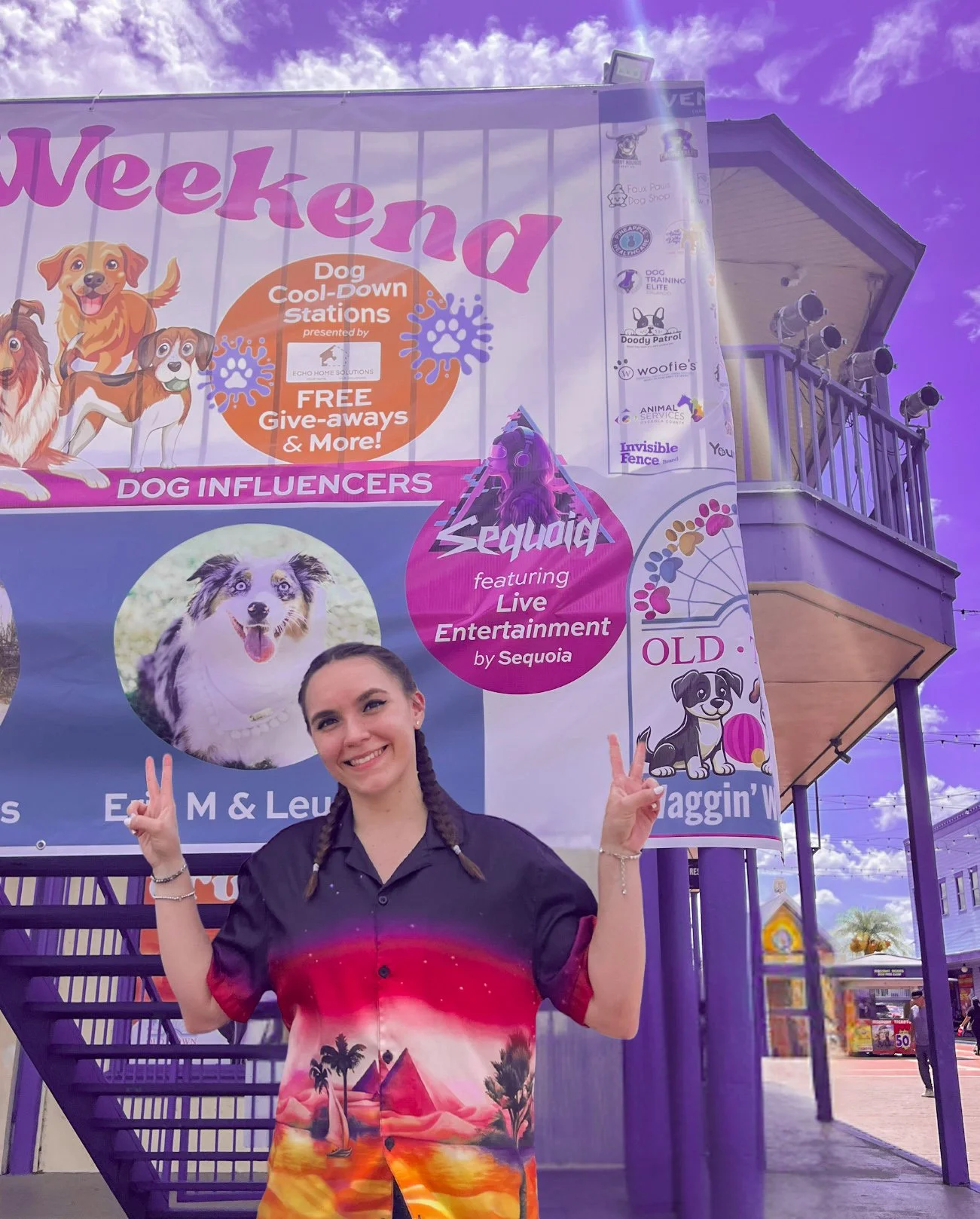 A young woman with braided hair smiling and making peace signs in front of a colorful event banner at a pet-themed event, with purple sky and event booths visible in the background.