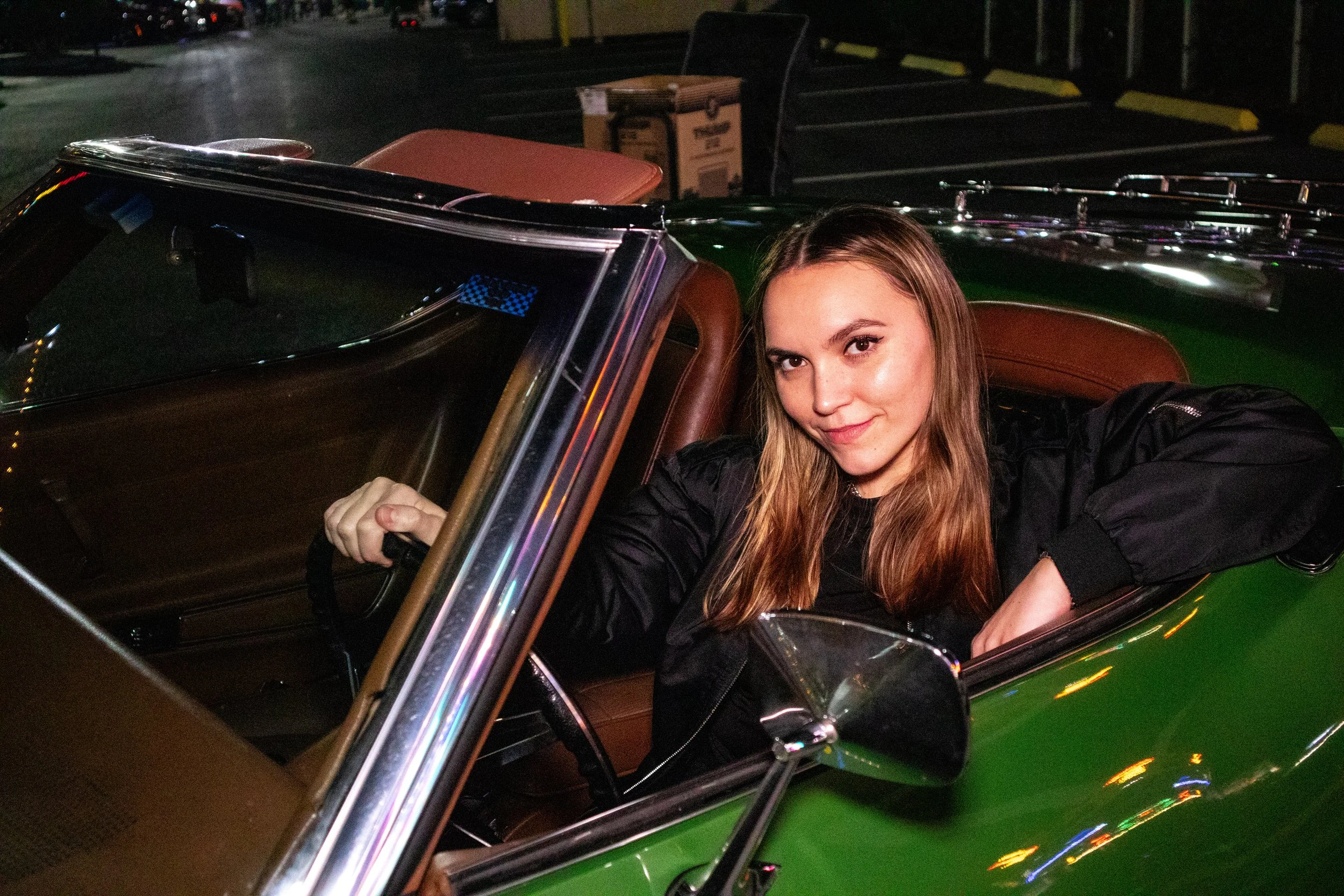 A young woman with long blonde hair smiling in a vintage green convertible car at night.
