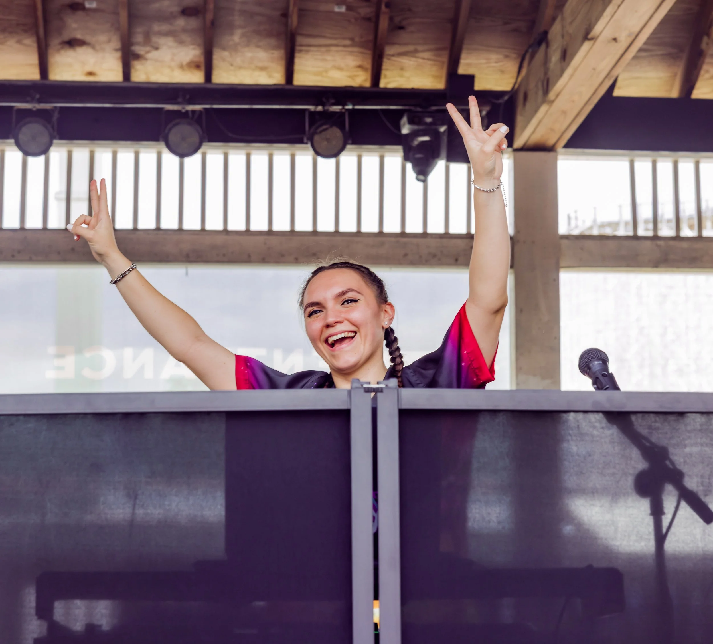 Person smiling and making peace signs with both hands inside a room with a high wooden ceiling, microphones, and a stage.