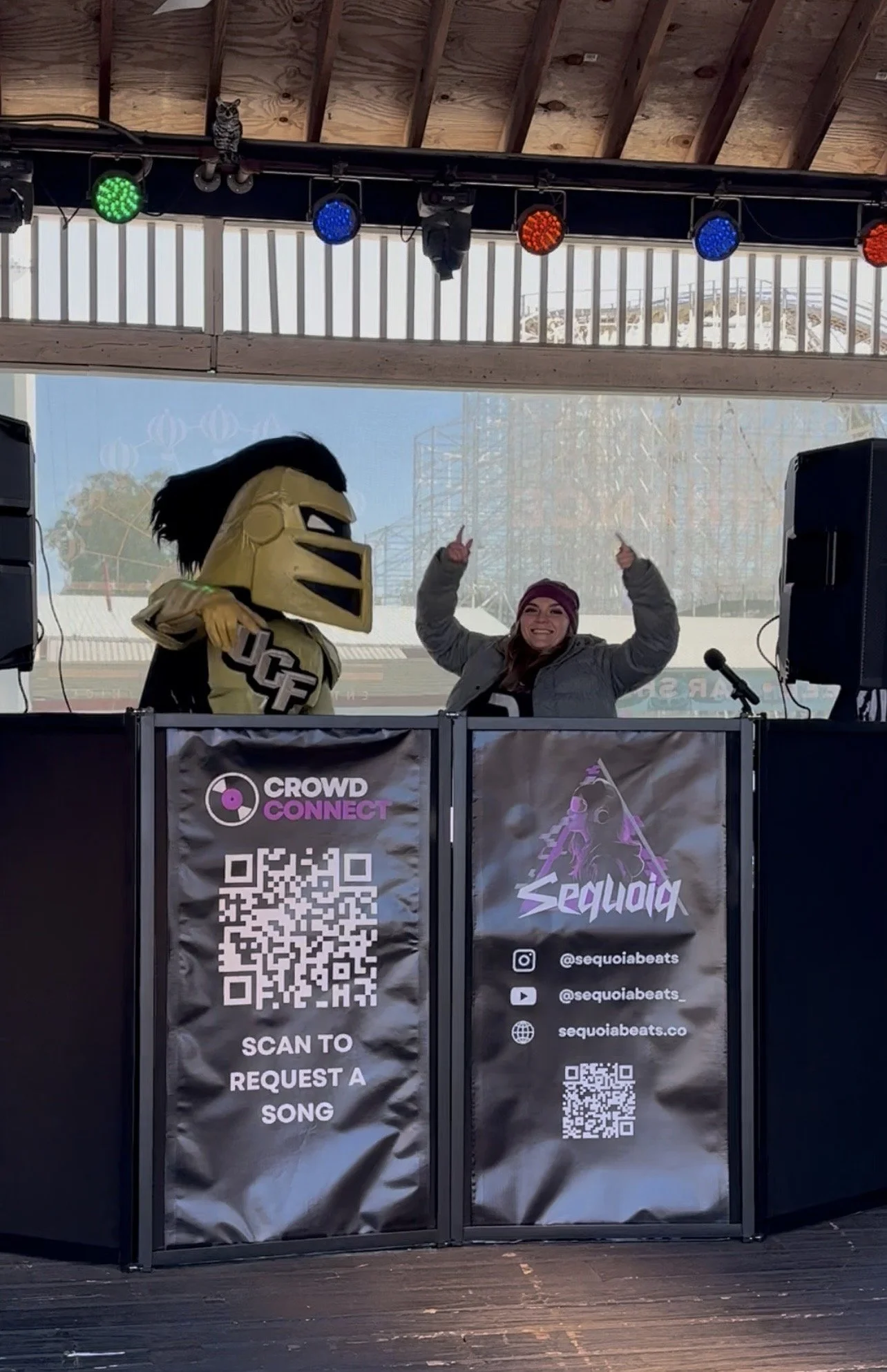 A woman and a person in a mascot costume are behind a DJ booth. The woman is smiling and raising her arms, wearing a beanie. The mascot has a yellow helmet with black hair and is wearing a racing jersey with the letters "UCF." The booth has promotion