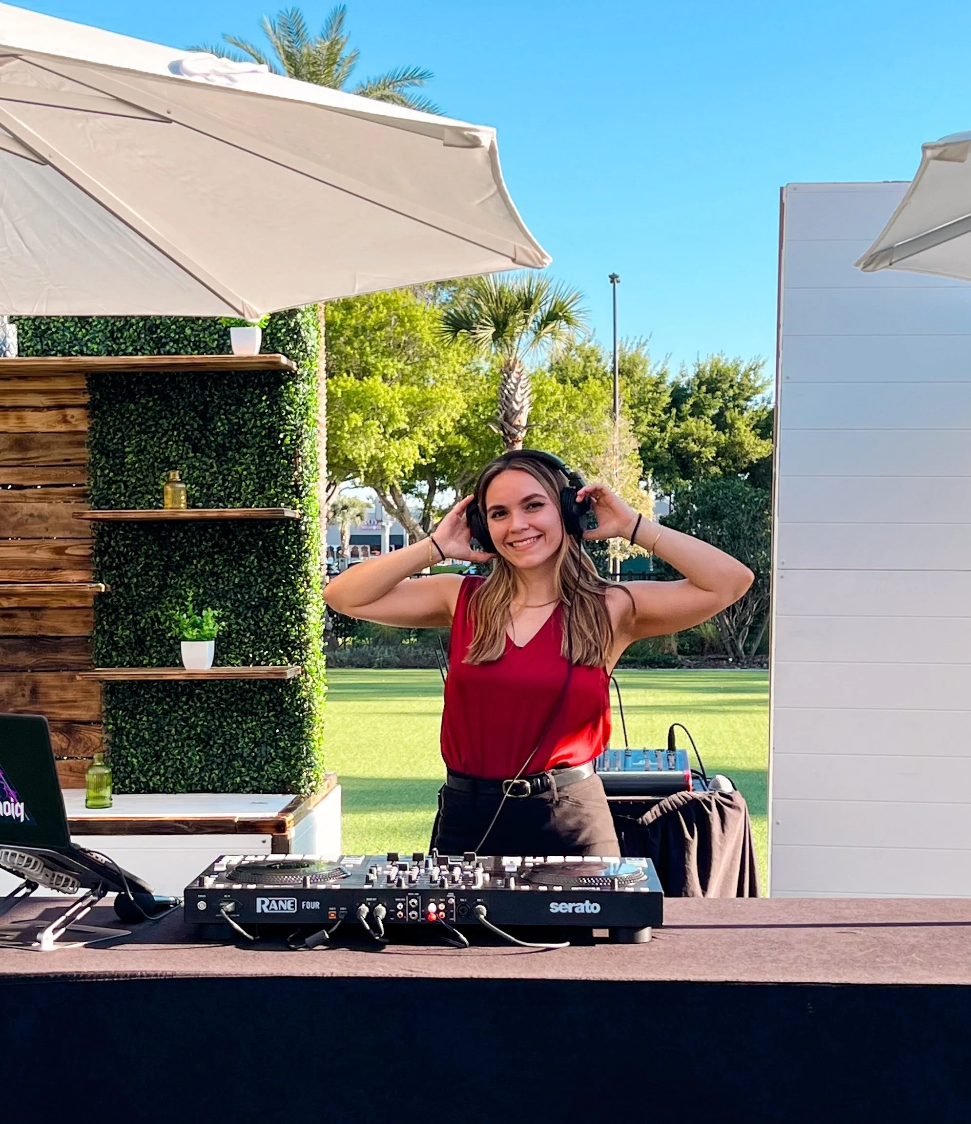 A young woman wearing a red top and black pants is smiling and adjusting headphones in front of DJ equipment outdoors. There are umbrellas, trees, and a clear blue sky in the background.
