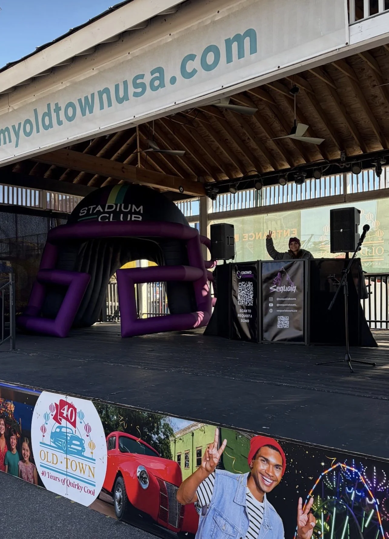 Outdoor stage with a woman waving, a large inflatable purple and black structure labeled 'Stadium Club,' and a banner advertising 'Old Town 40 Years of Quirky Cool' with a smiling man posing with a peace sign and a Ferris wheel in the background.