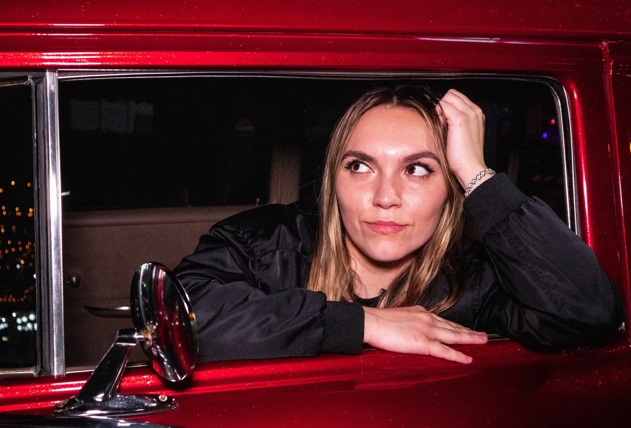 A young woman with long blonde hair, wearing a black jacket and a bracelet, rests her arms on a red car window frame while looking to her left at nighttime. The car's side mirror and doorframe are visible.
