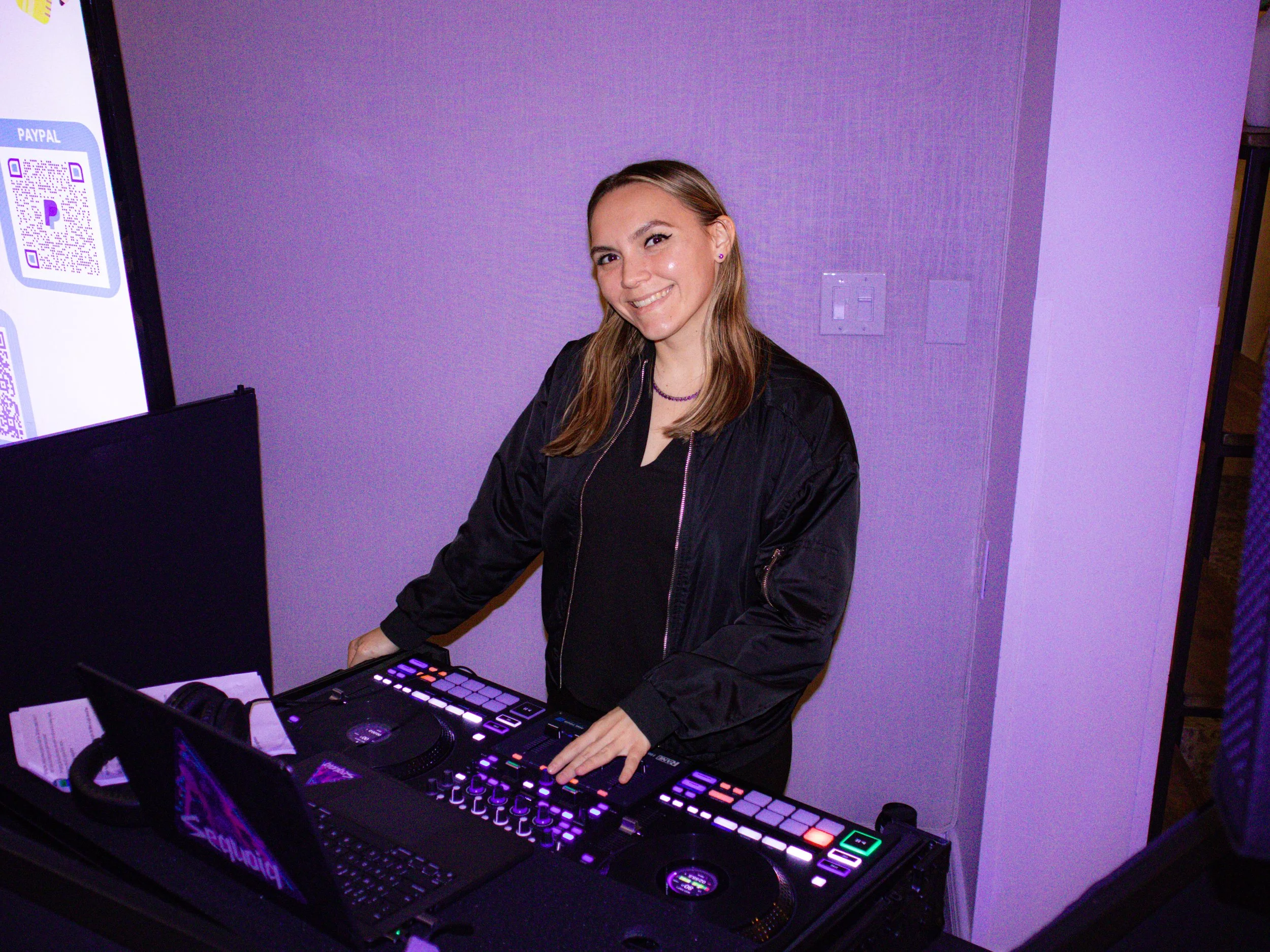 A woman with blonde hair smiling at a DJ setup with colorful LED lights, in a purple-lit room.