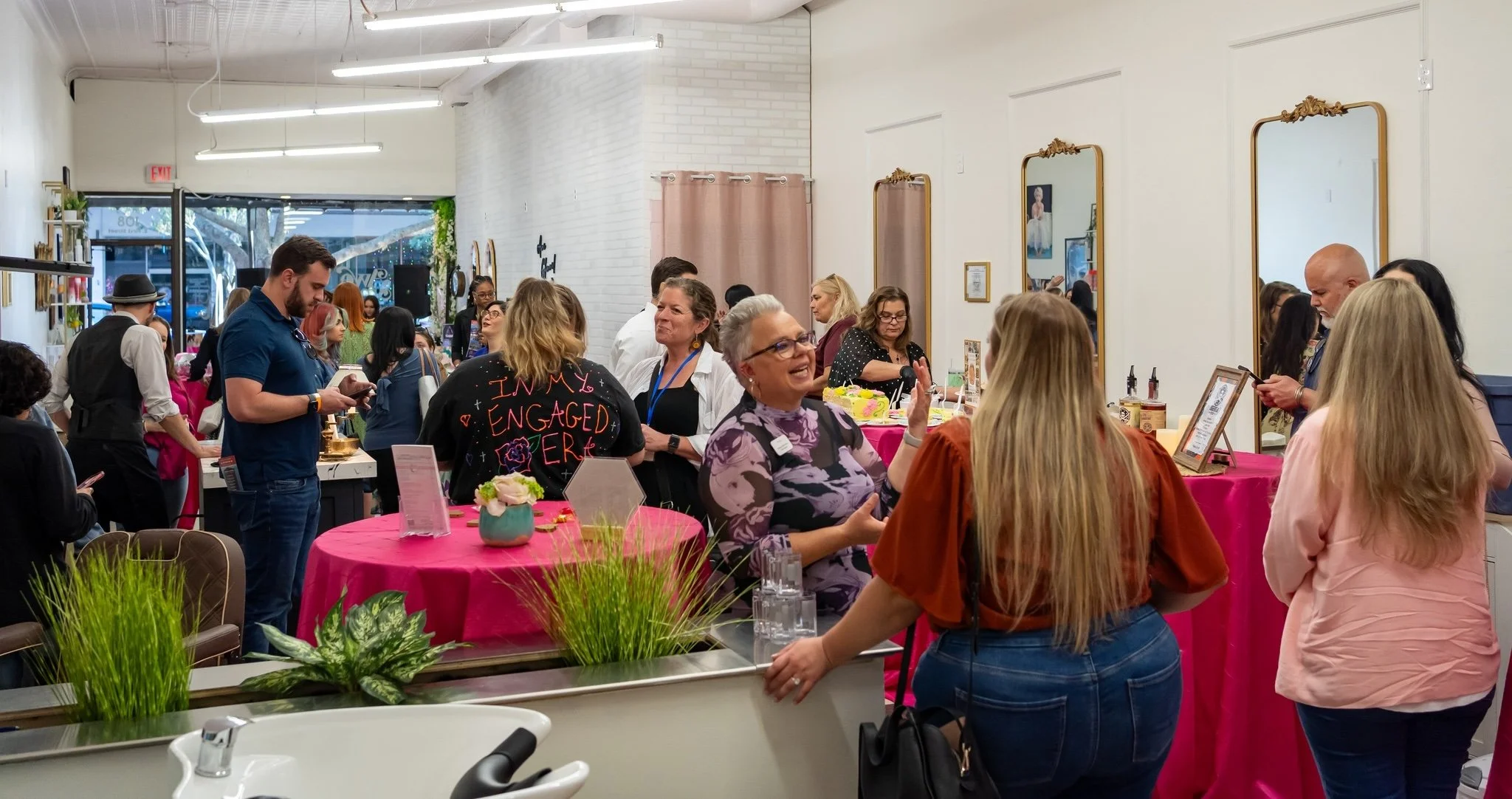 A lively indoor event with people socializing around pink-covered tables decorated with flowers, in a well-lit room with white walls, mirrors, and a large window showing trees outside.
