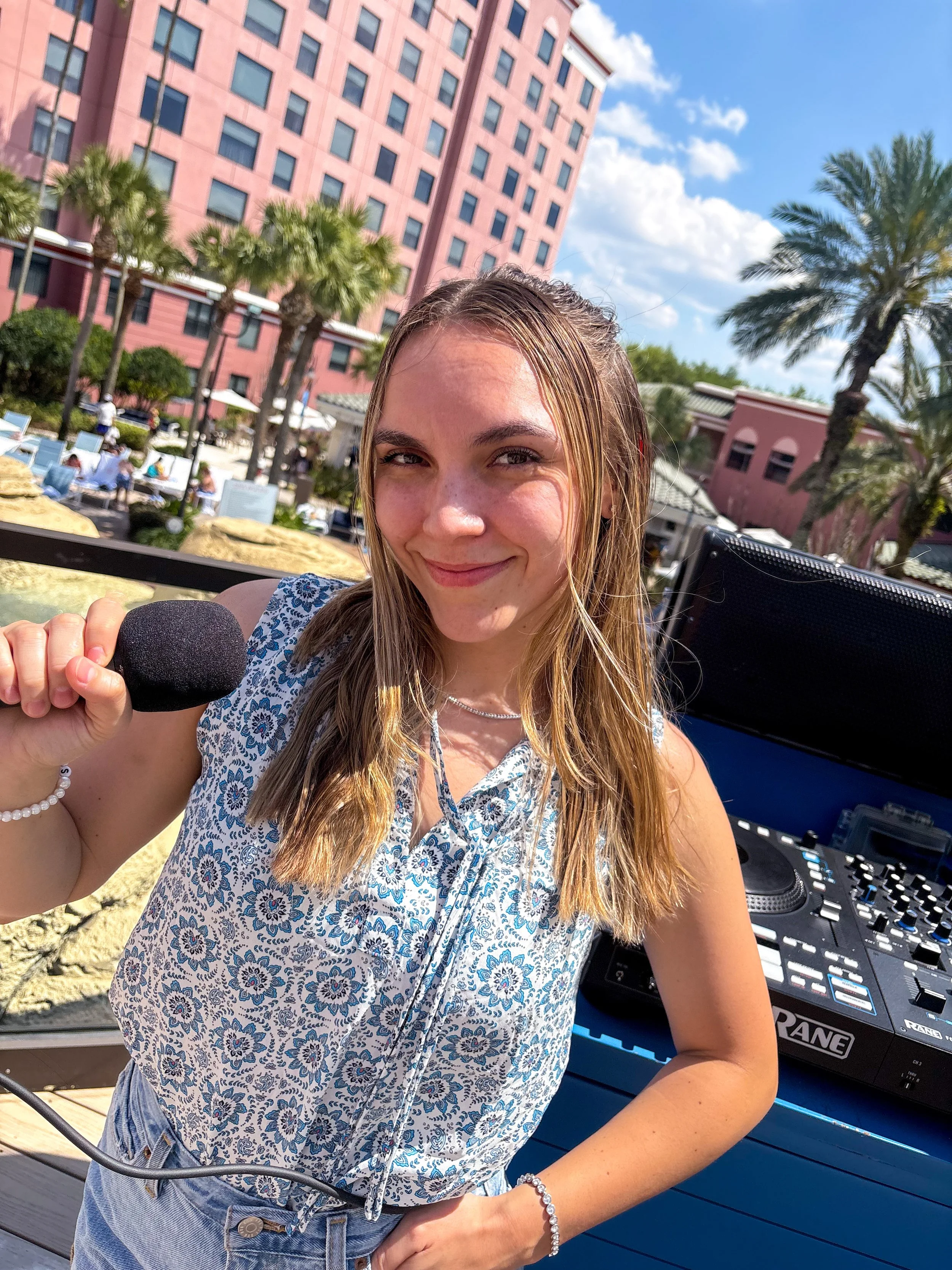 A young woman is smiling at the camera while holding a microphone. She is standing outdoors near a DJ booth with a mixer, with a pink building, palm trees, and a blue sky in the background.