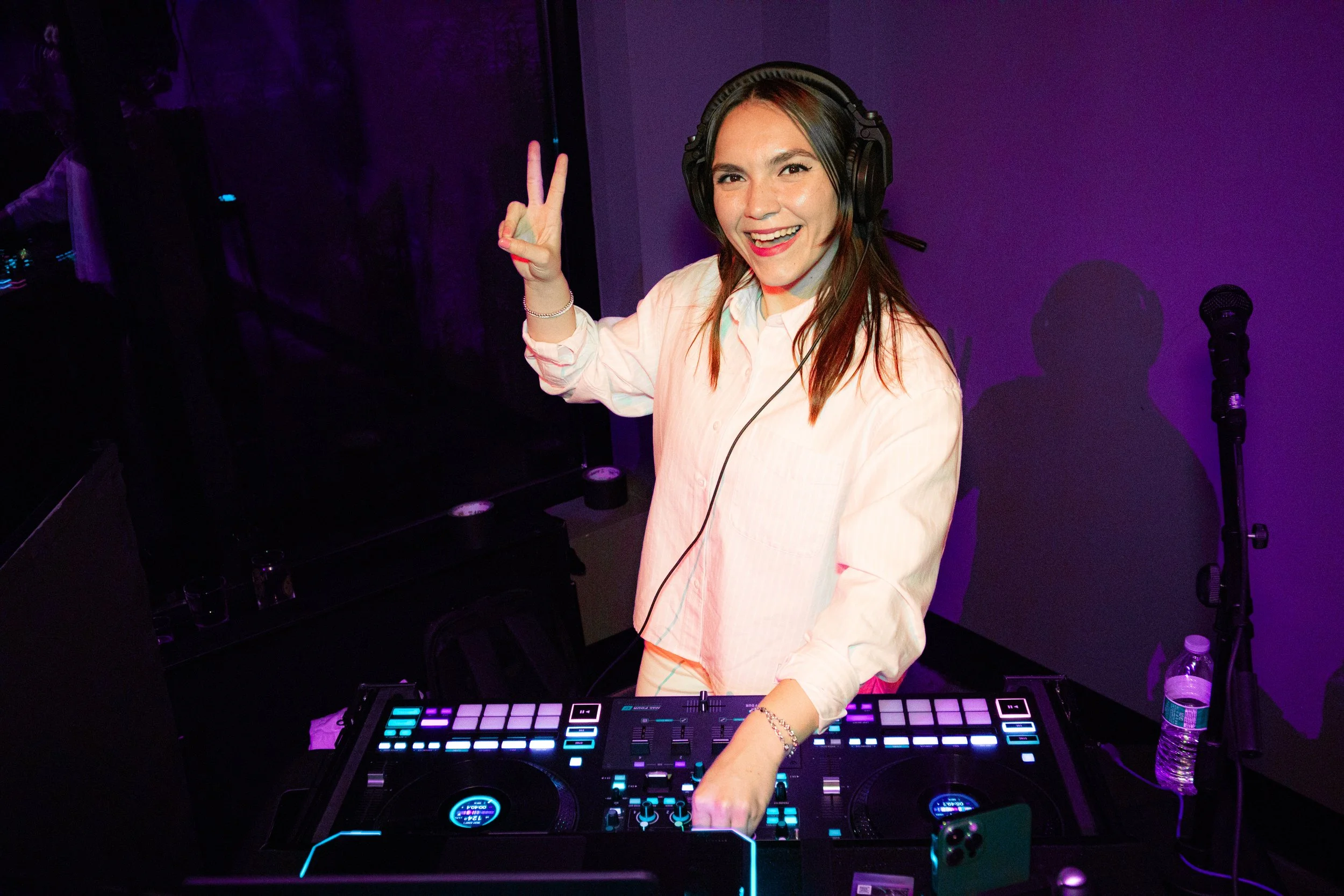 A woman with a headset and a big smile is DJing at a nightclub, making a peace sign with her right hand, in front of DJ equipment with colorful LED buttons, purple lighting, and a microphone on a stand nearby.