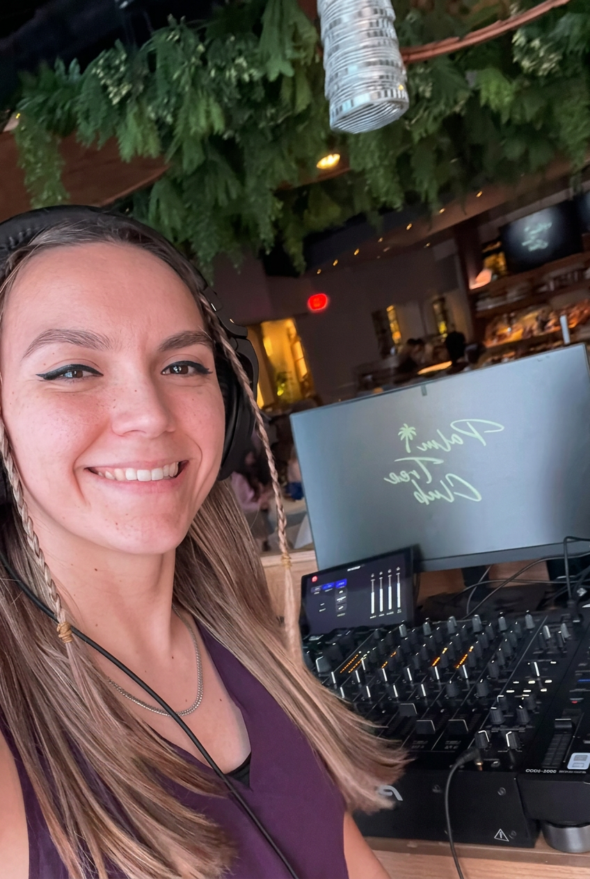 A smiling woman with long blonde hair and freckles, wearing a purple top and headphones, stands in front of DJ equipment with a computer screen displaying 'Paradise Club Tree'. The background shows a lively indoor setting with greenery, a bar, and people.