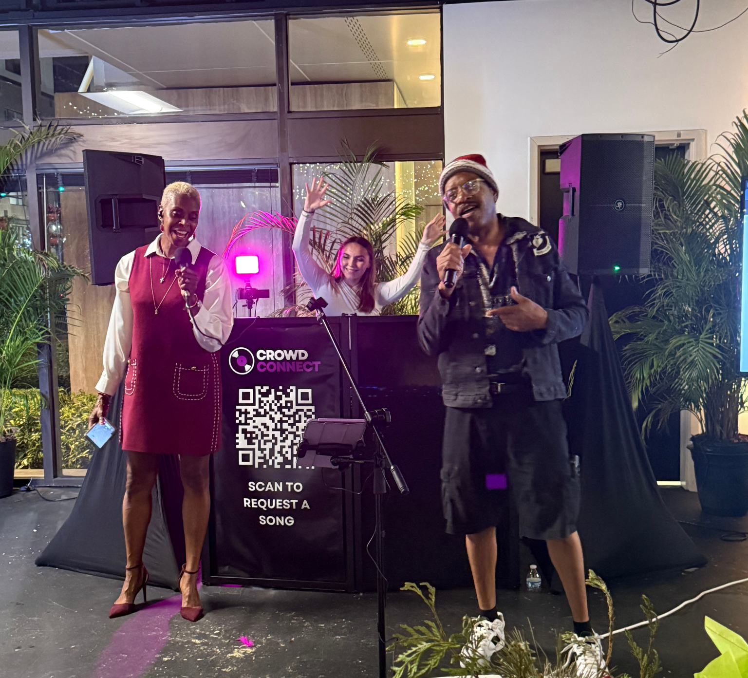 People performing karaoke at a night event, with two women holding microphones and a man wearing a Santa hat singing, supported by plants and a sign with a QR code for song requests.