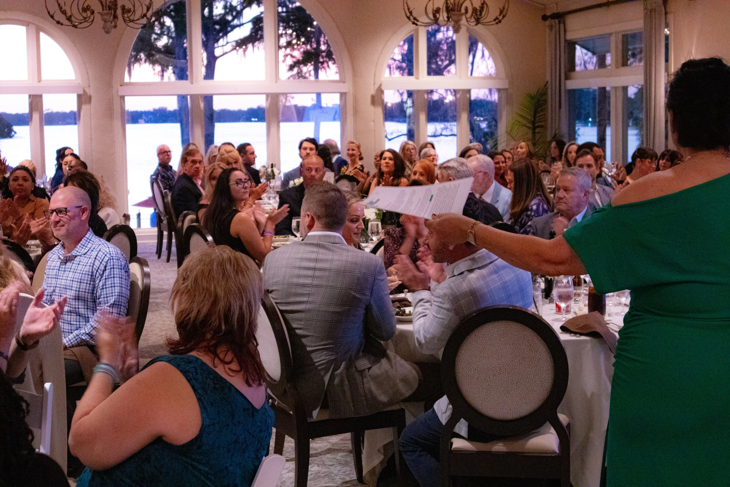 A large group of people sitting at banquet tables inside a well-lit room with large windows. The people are clapping and paying attention to a person standing and speaking while holding a piece of paper. The scene appears to be a formal or celebrator