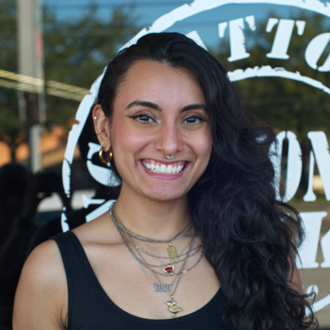 A smiling woman with black, wavy hair and multiple layered necklaces stands outdoors in front of a window with lettering.