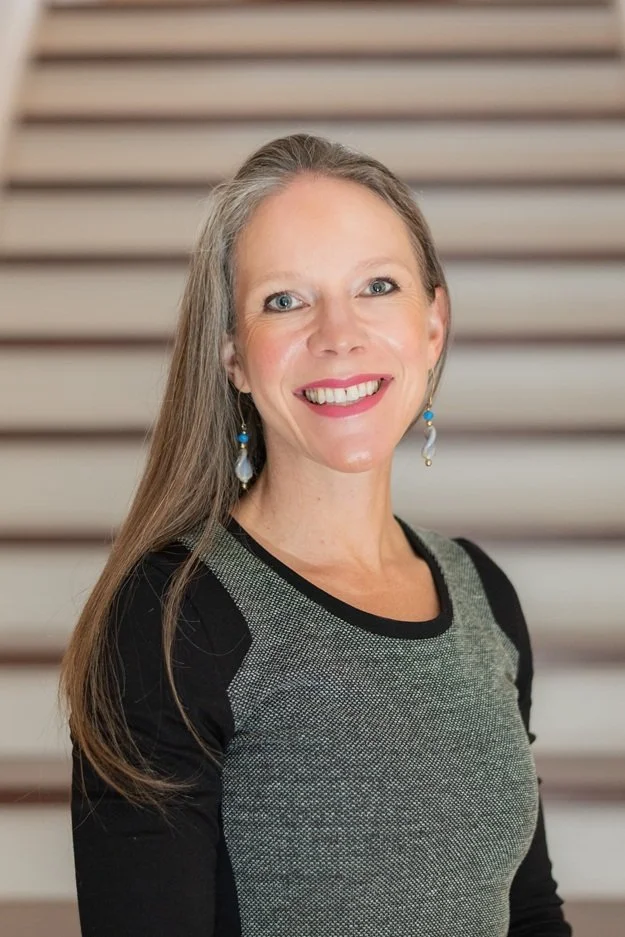 A portrait of a middle-aged woman with long gray hair, blue eyes, and earrings, standing in front of a staircase with wooden steps and a white railing. She is smiling and wearing a gray and black top.