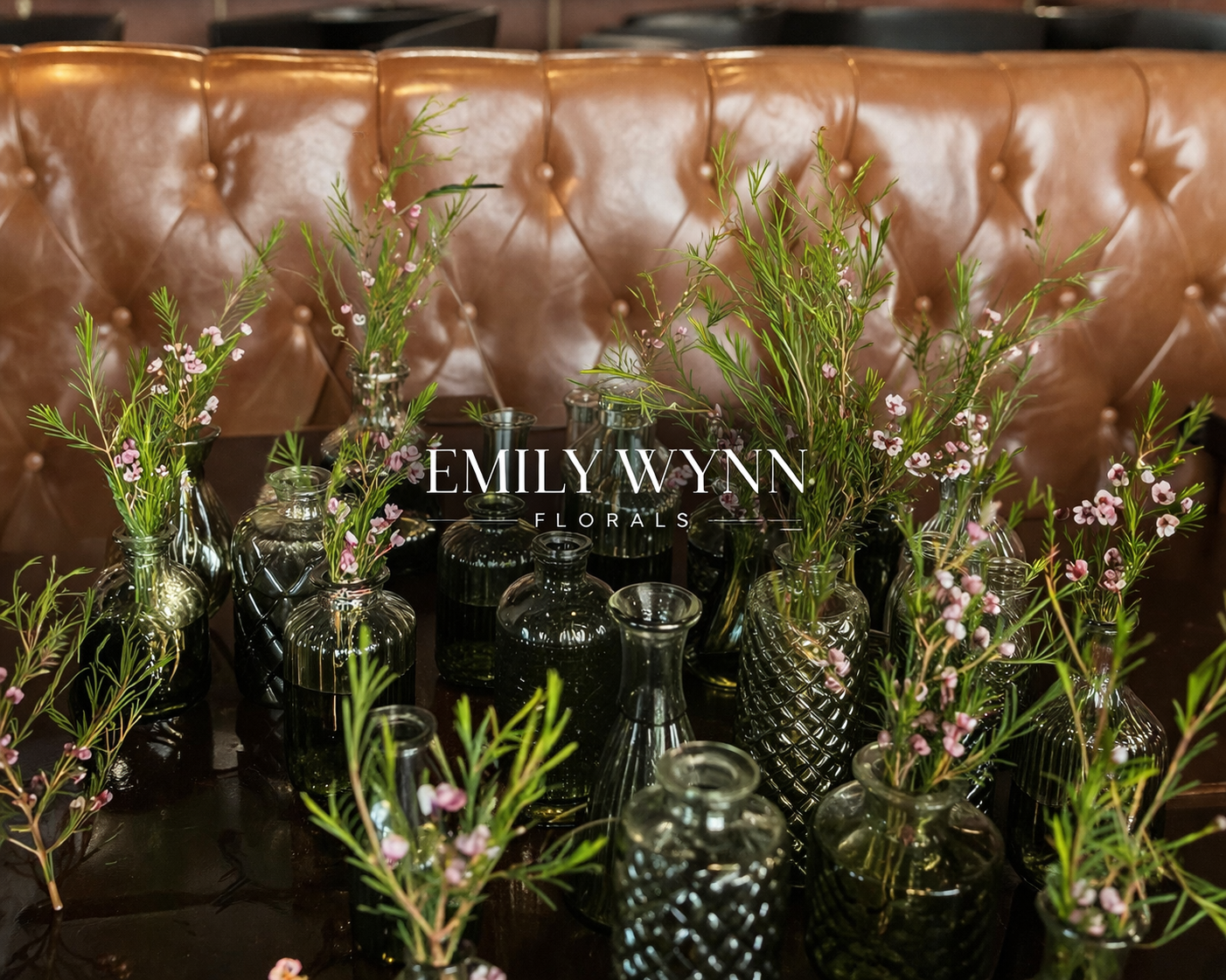 Decorative arrangement of small pink and green flowers in dark glass vases on a polished dark table, with a brown leather tufted bench in the background.