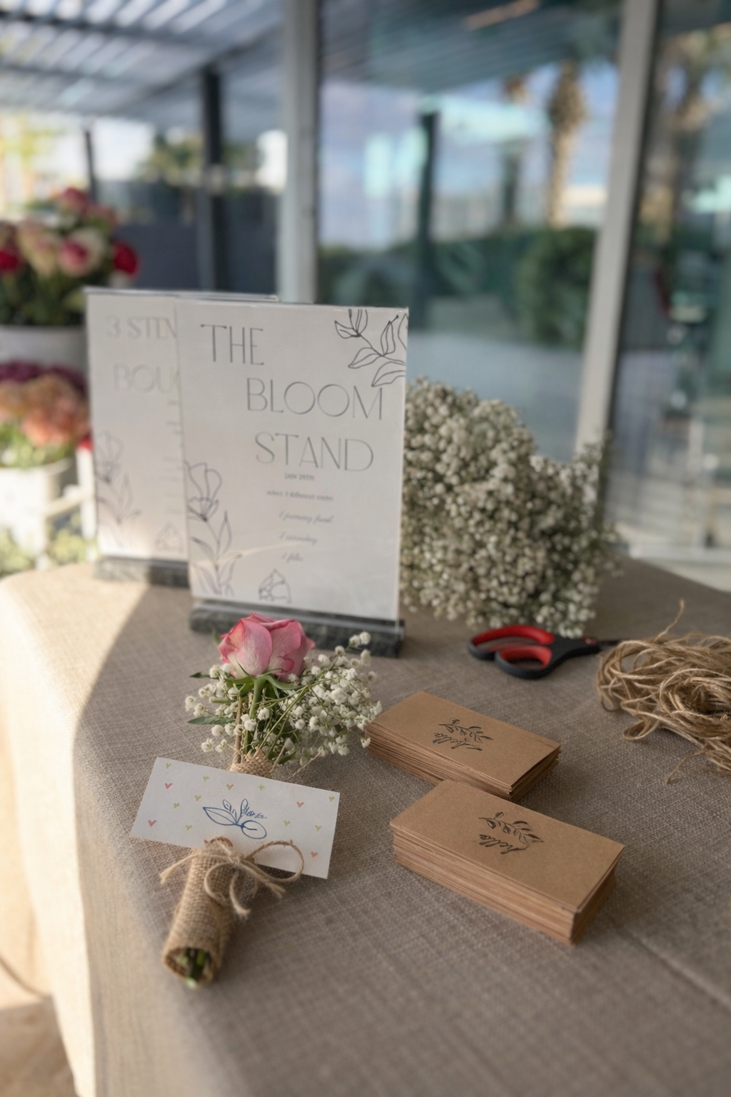 A table decorated with flowers, a sign reading 'The Bloom Stand', stacks of brown tags with a floral design, a pair of scissors, and a spool of twine, set in a bright outdoor space.