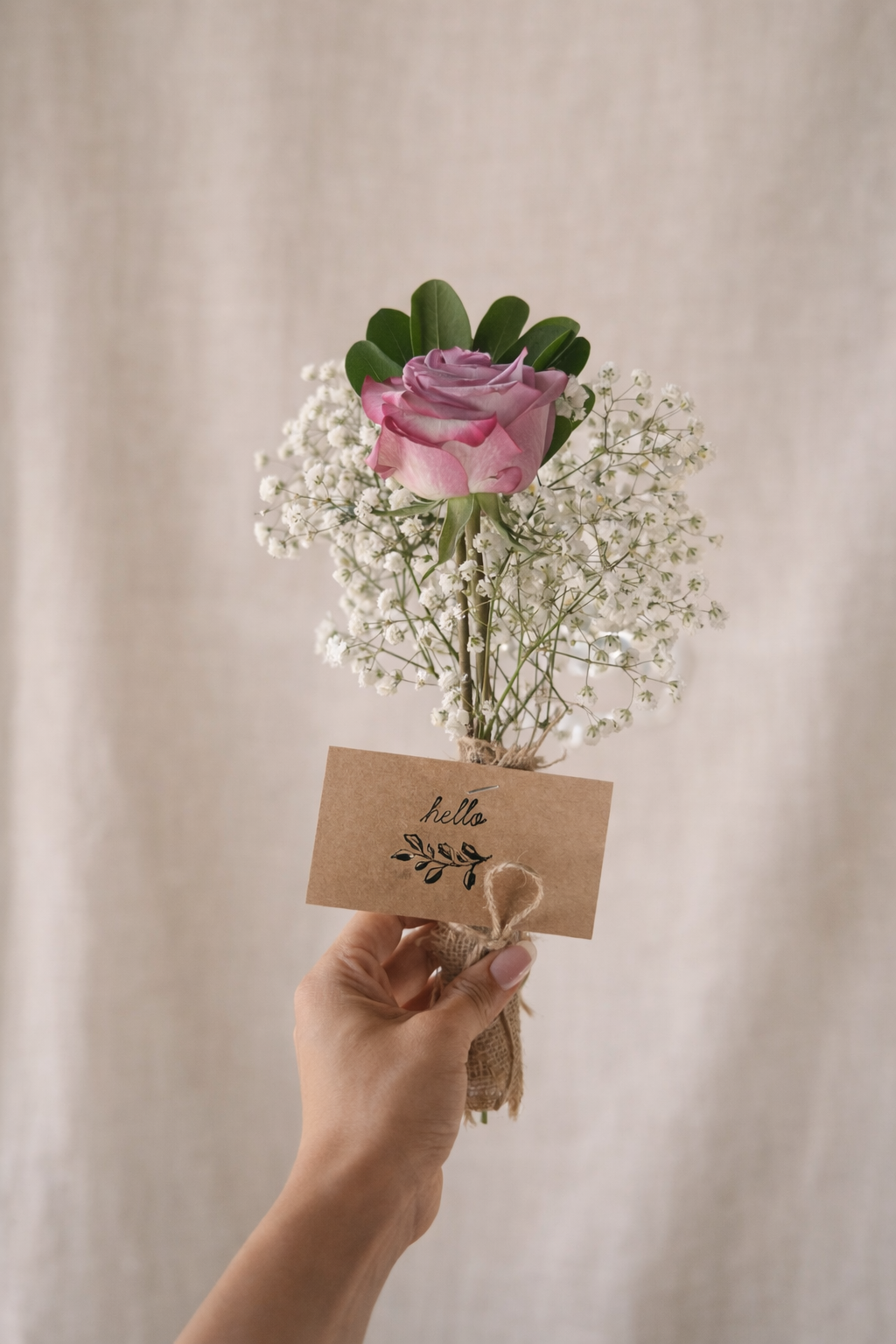 Hand holding a small bouquet of pink rose, white baby's breath, and green leaves with a "hello" note tied to the bouquet.
