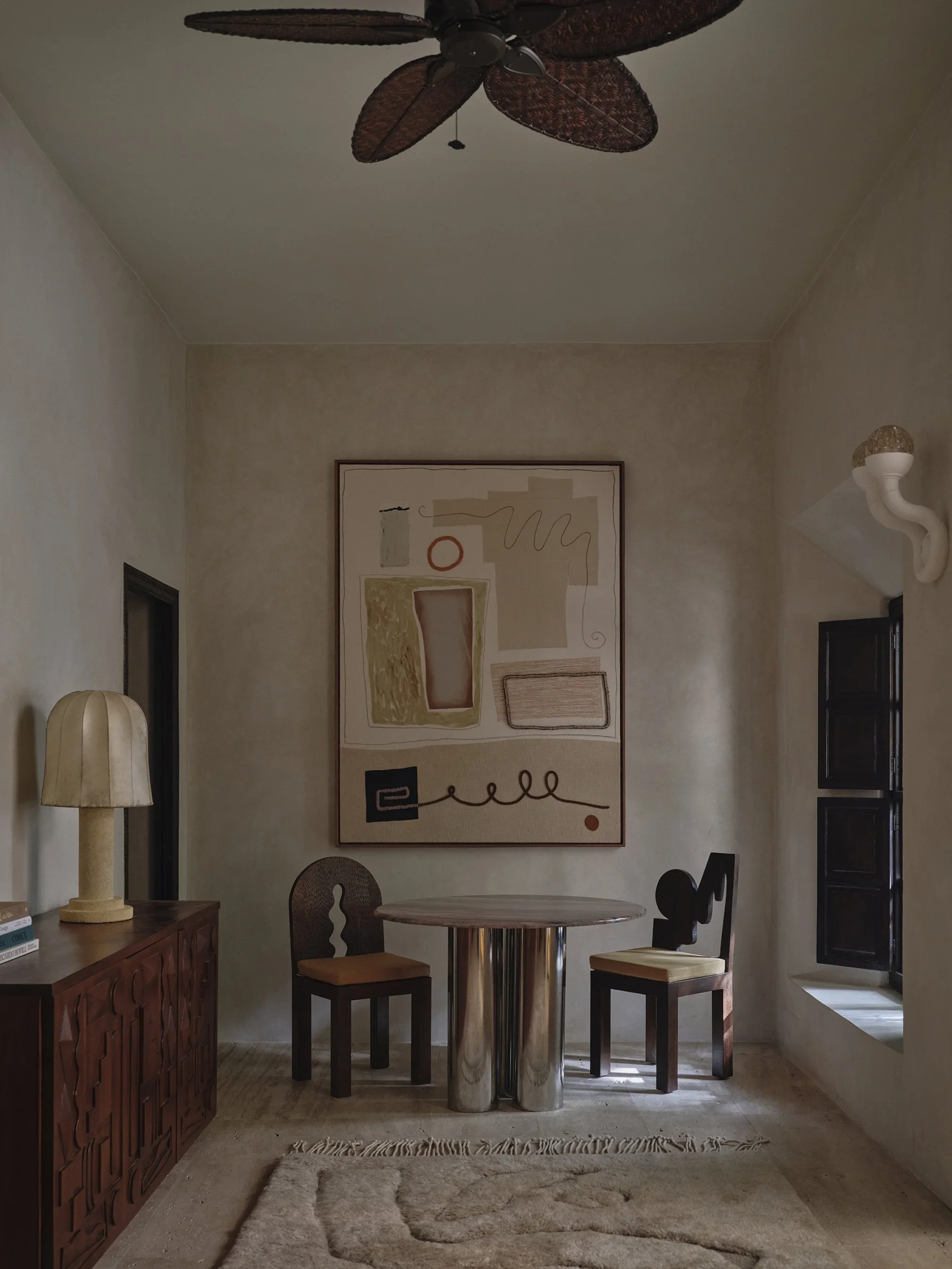 A cozy corner with a small round table with a metallic base, two wooden chairs with upholstered seats, a wooden sideboard with books and a lamp, a large abstract artwork on the wall, a textured rug on the floor, and a window with black shutters letting in natural light.