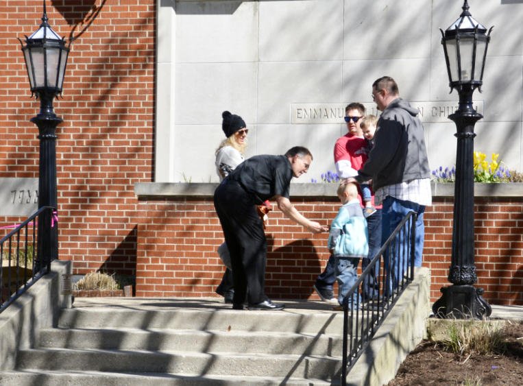 A group of people, including children and adults, gathered outside a brick building with a wall memorial, on steps with black railings, with two black lampposts on either side.