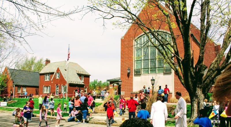 People gather outside a red brick church with large windows and a tall steeple, surrounded by trees and a rural neighborhood, on a cloudy day.