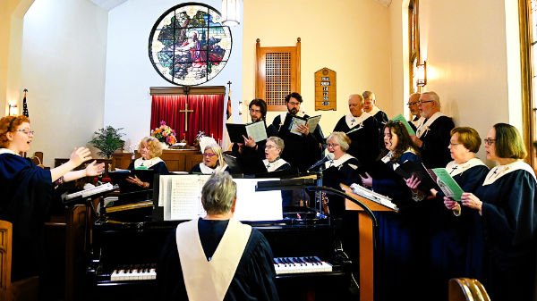 A choir of adults singing and a conductor leading them in a church, with a pianist playing a black grand piano.