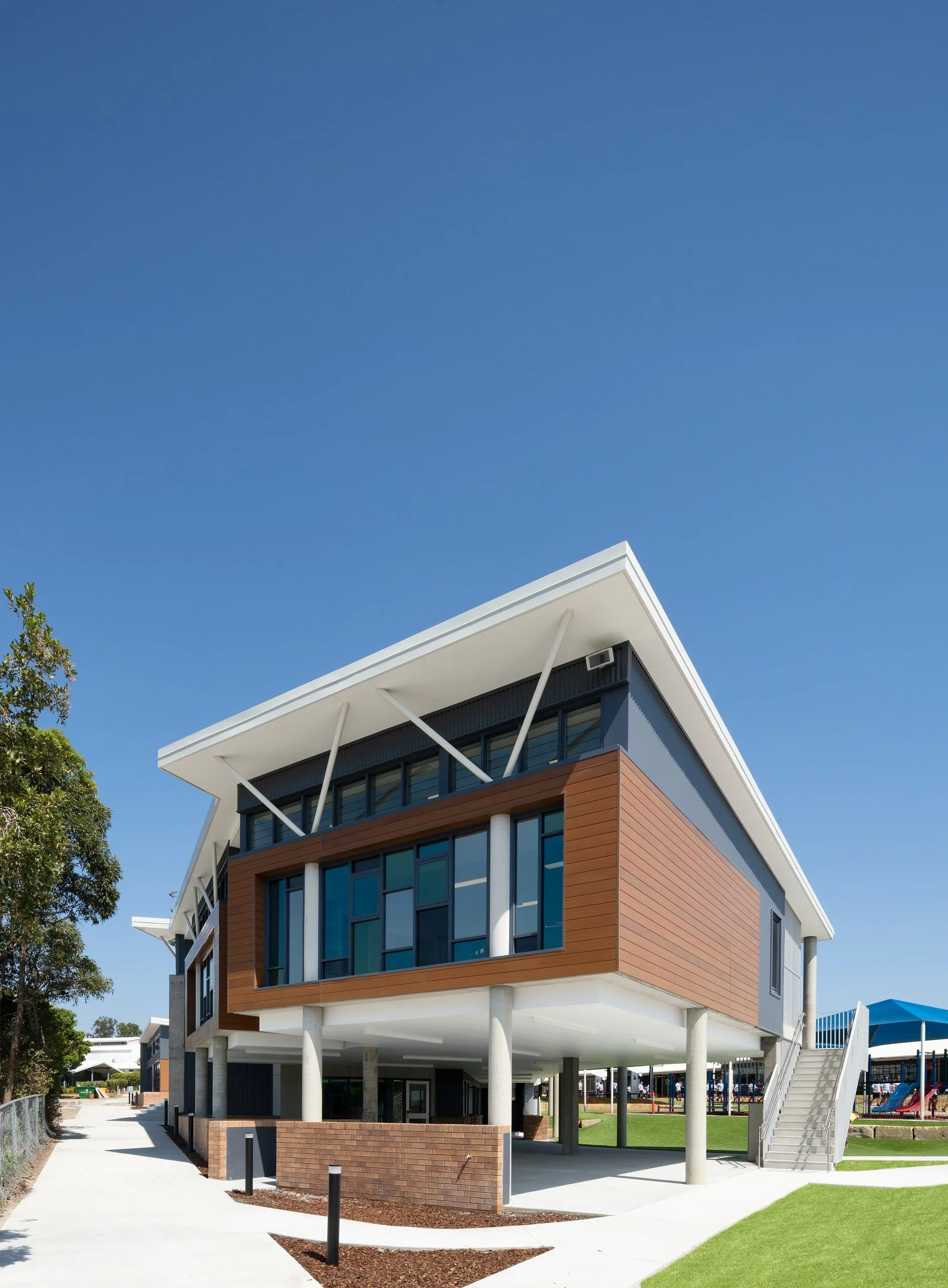 Modern two-story building with large windows, white structural supports, and brown exterior siding, set against a clear blue sky.