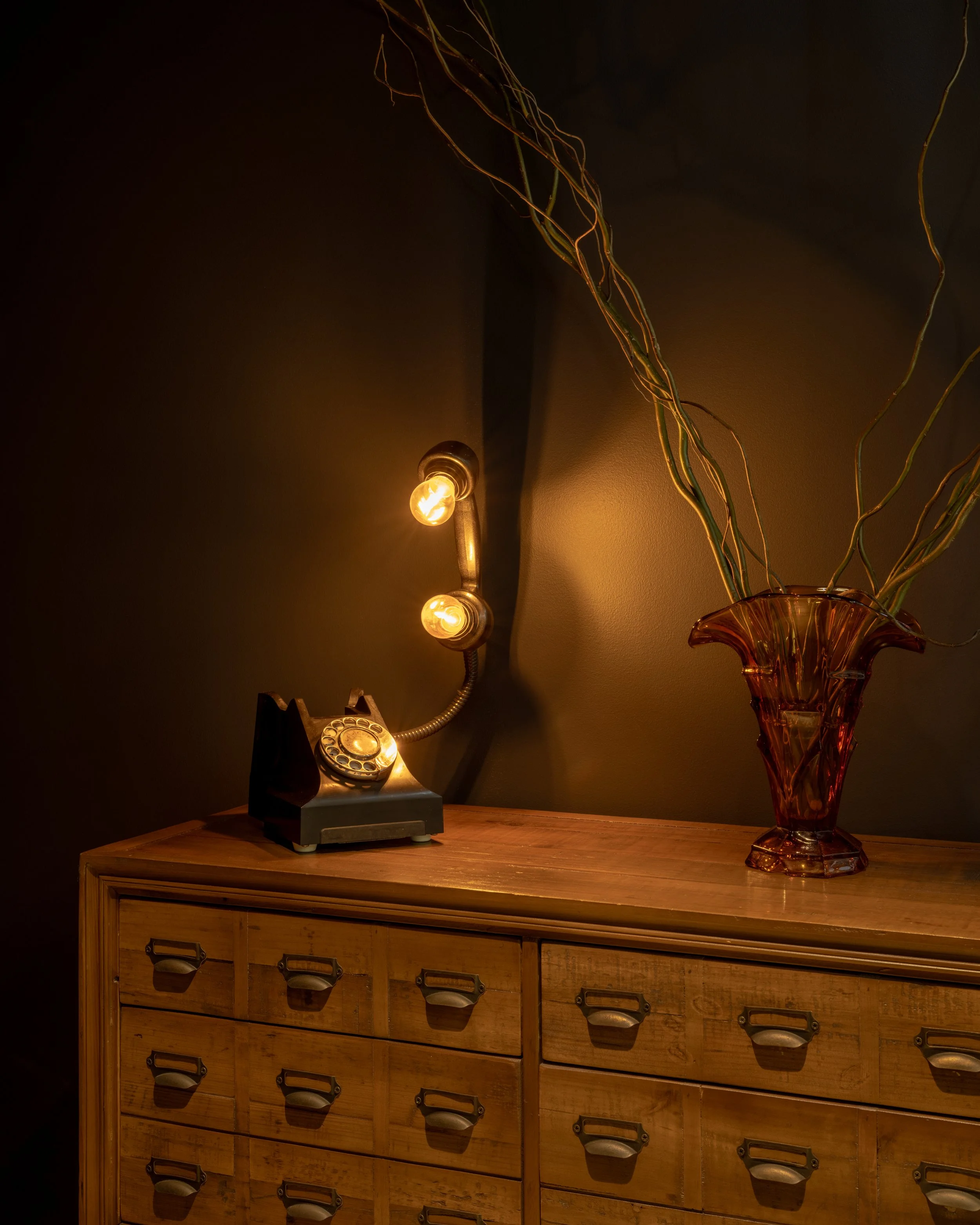 A vintage rotary phone on a wooden dresser next to a large amber glass vase with decorative twisted branches inside, illuminated by a wall-mounted light fixture with two exposed bulbs.