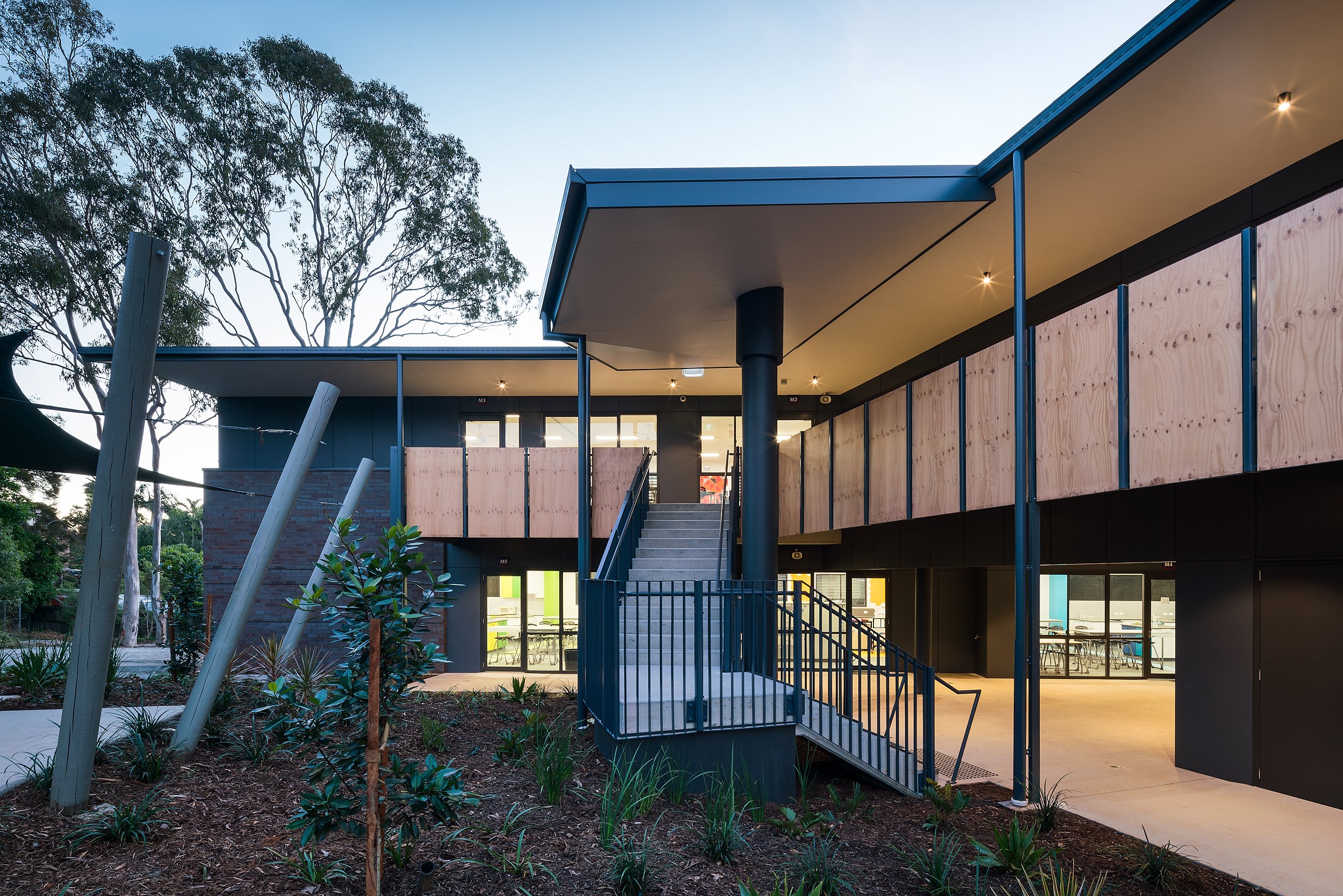 Modern building with black exterior, large windows, and a staircase leading to an upper level, surrounded by trees and landscaped plants.