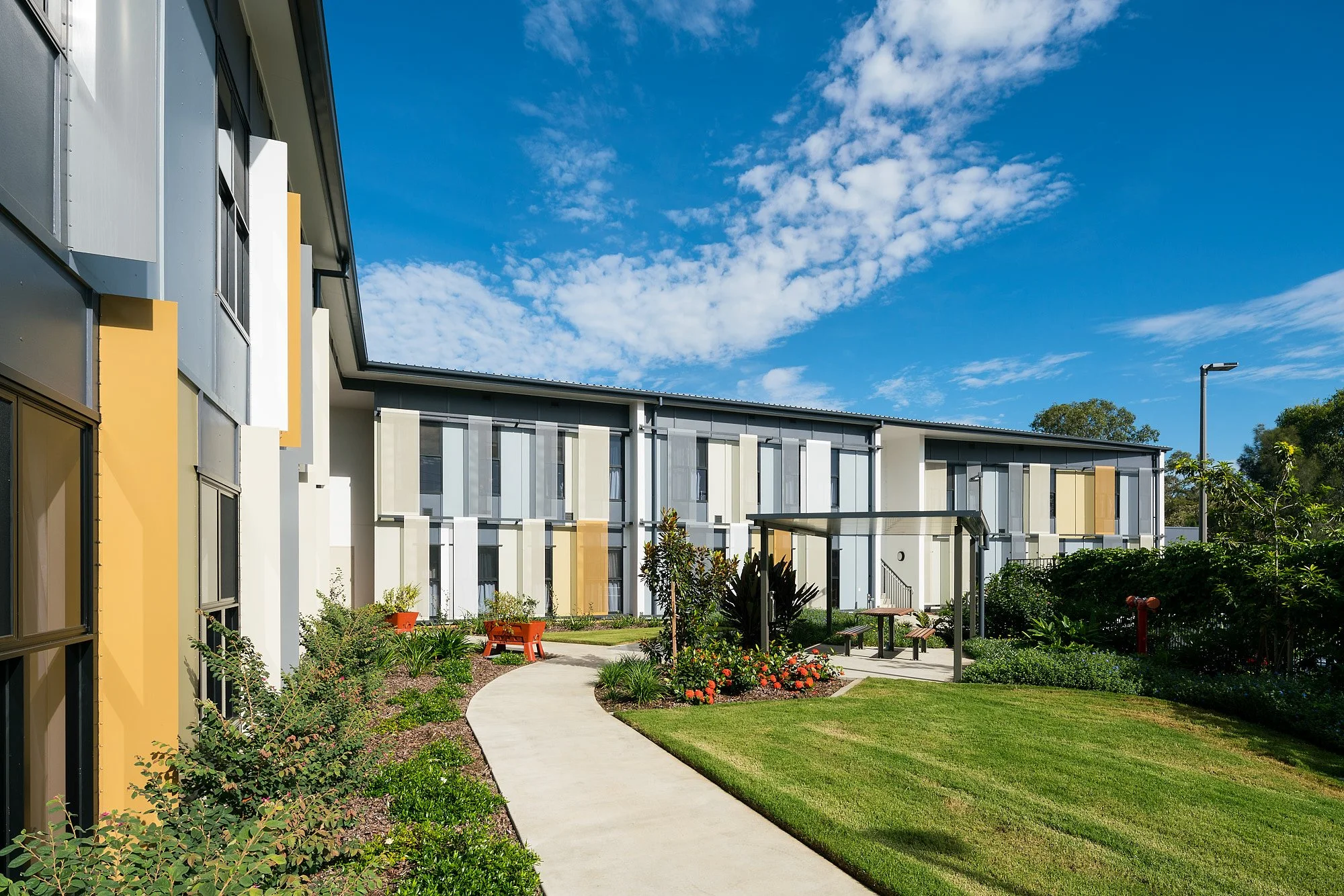 Modern apartment complex with landscaped courtyard, walking path, garden beds, benches, and a covered seating area under a bright blue sky.