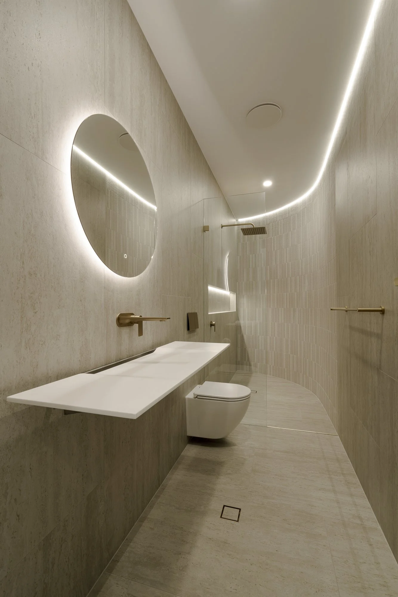 Minimalist bathroom with curved beige walls, a white countertop, a round mirror, a wall-mounted toilet, and a ceiling-mounted shower head with a glass shower enclosure.