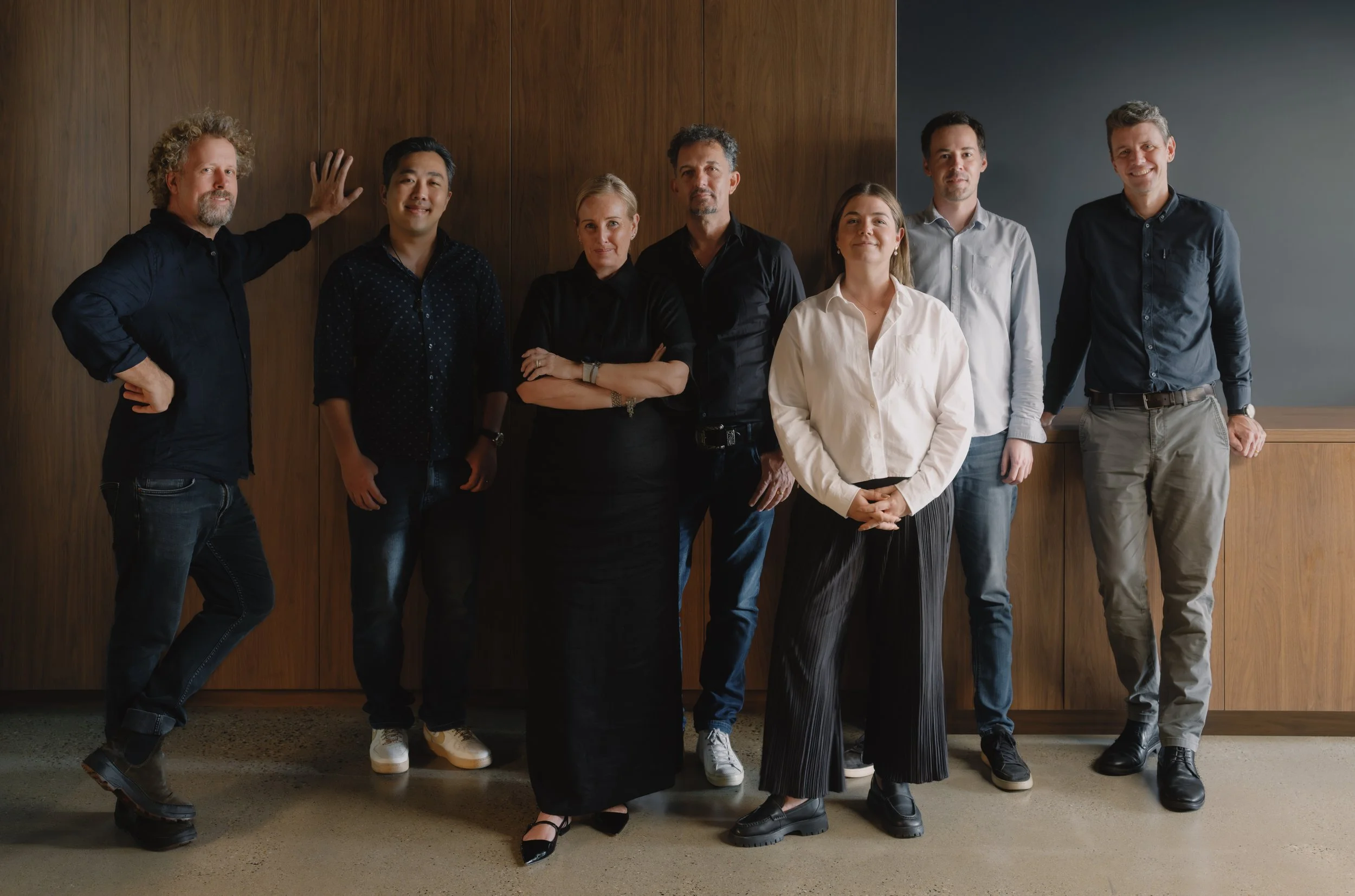 Group of seven diverse professionals standing indoors against a wooden wall, posing for a photo.