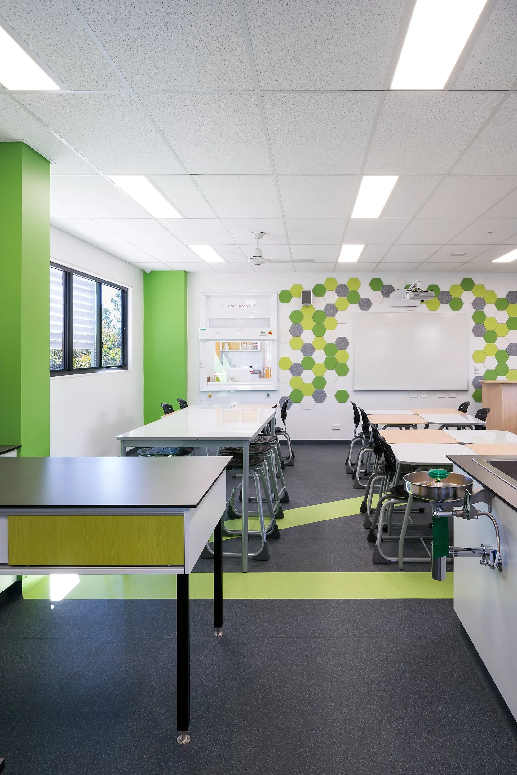 A modern classroom with green accents, hexagonal wall decorations, tables and chairs, and a whiteboard.
