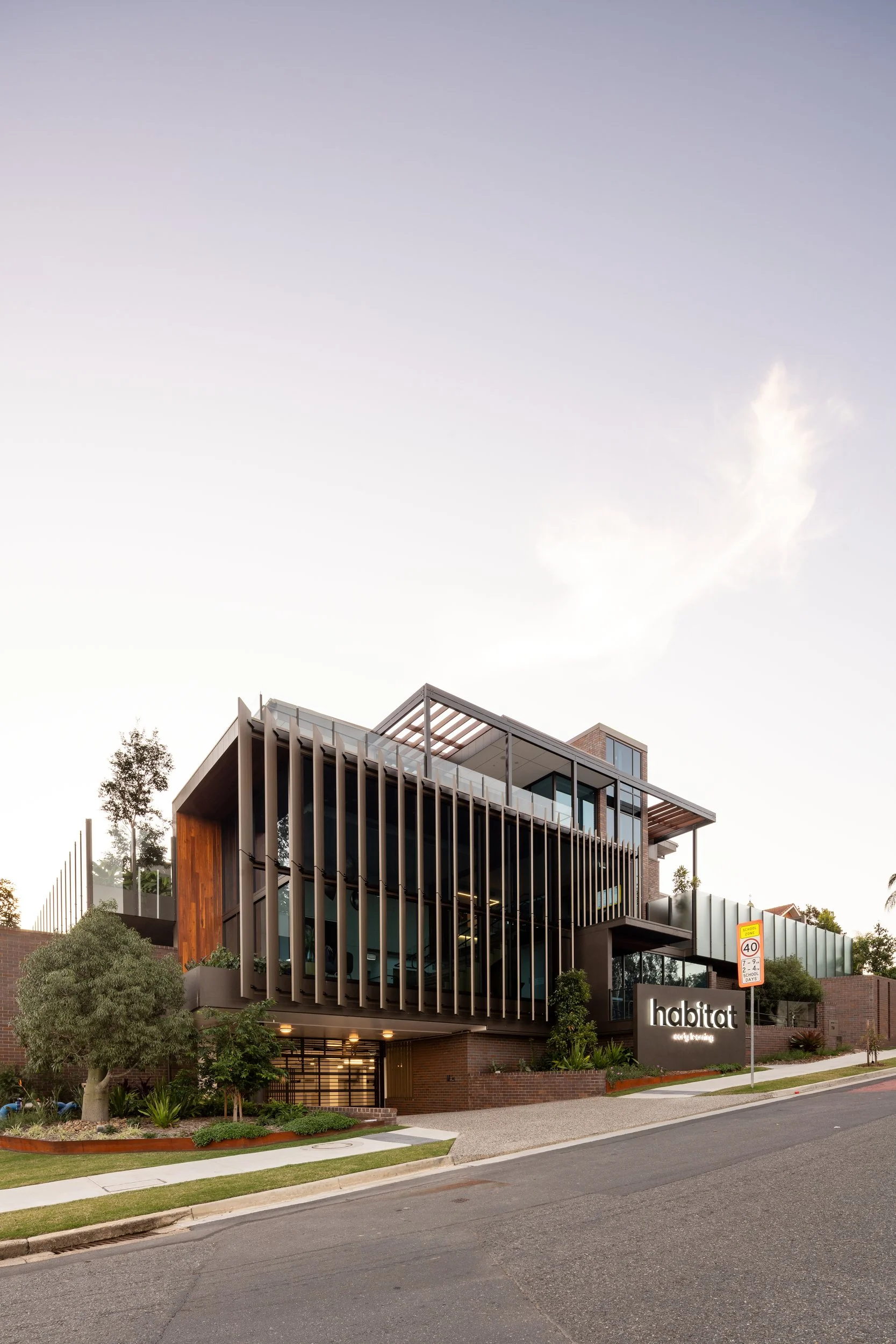 Modern multi-story building with wooden and metal architectural elements, landscaped area with trees and shrubs, sign that reads 'habitat early learning', and a street with a speed limit sign of 40 mph.