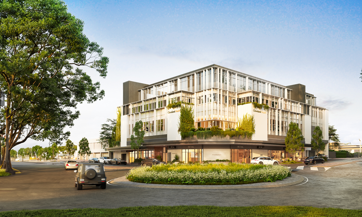Modern multi-story building with retail shops on the ground floor and residential or office units above, surrounded by trees and a roundabout with a landscaped island, under a clear blue sky.