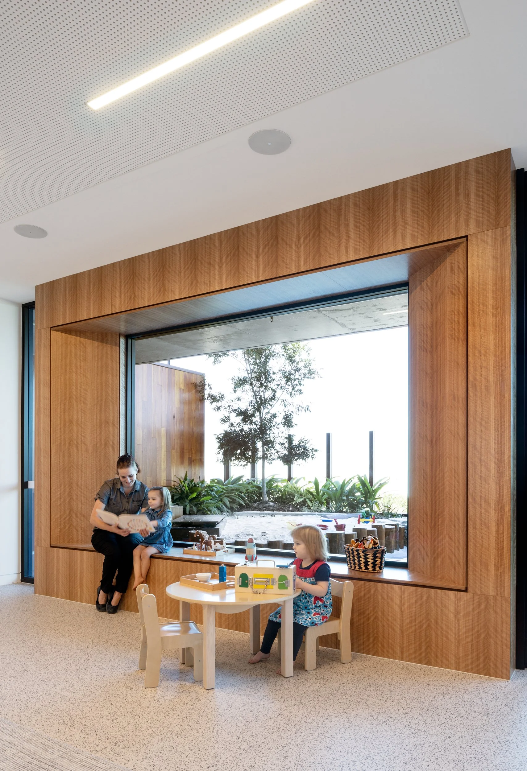 A woman and a young girl are sitting on a wooden window seat reading a book, while another young girl is sitting at a small table with toys inside an indoor play area with floor carpeting, large window, and outdoor garden with plants and trees.