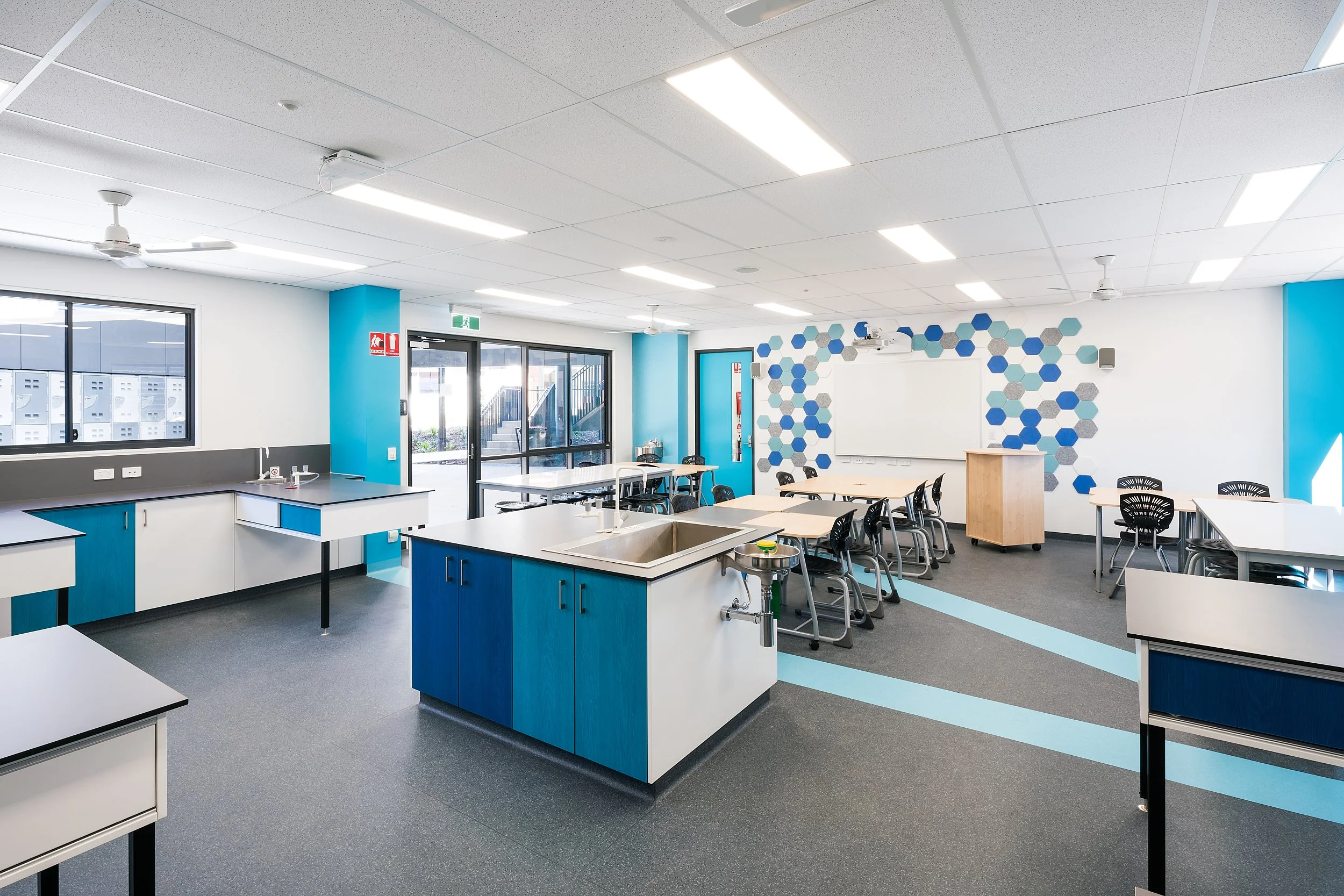 Bright classroom with blue, white, and gray decor, tables and chairs arranged for group work, a whiteboard at the front, and a science sink station, with windows and doors to the outside.