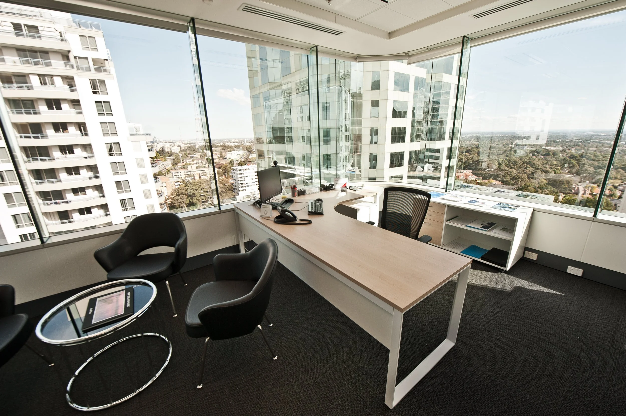 Modern office with a large wooden desk, multiple chairs, a computer, phone, and office supplies, surrounded by floor-to-ceiling windows with a cityscape view of tall buildings and clear sky.