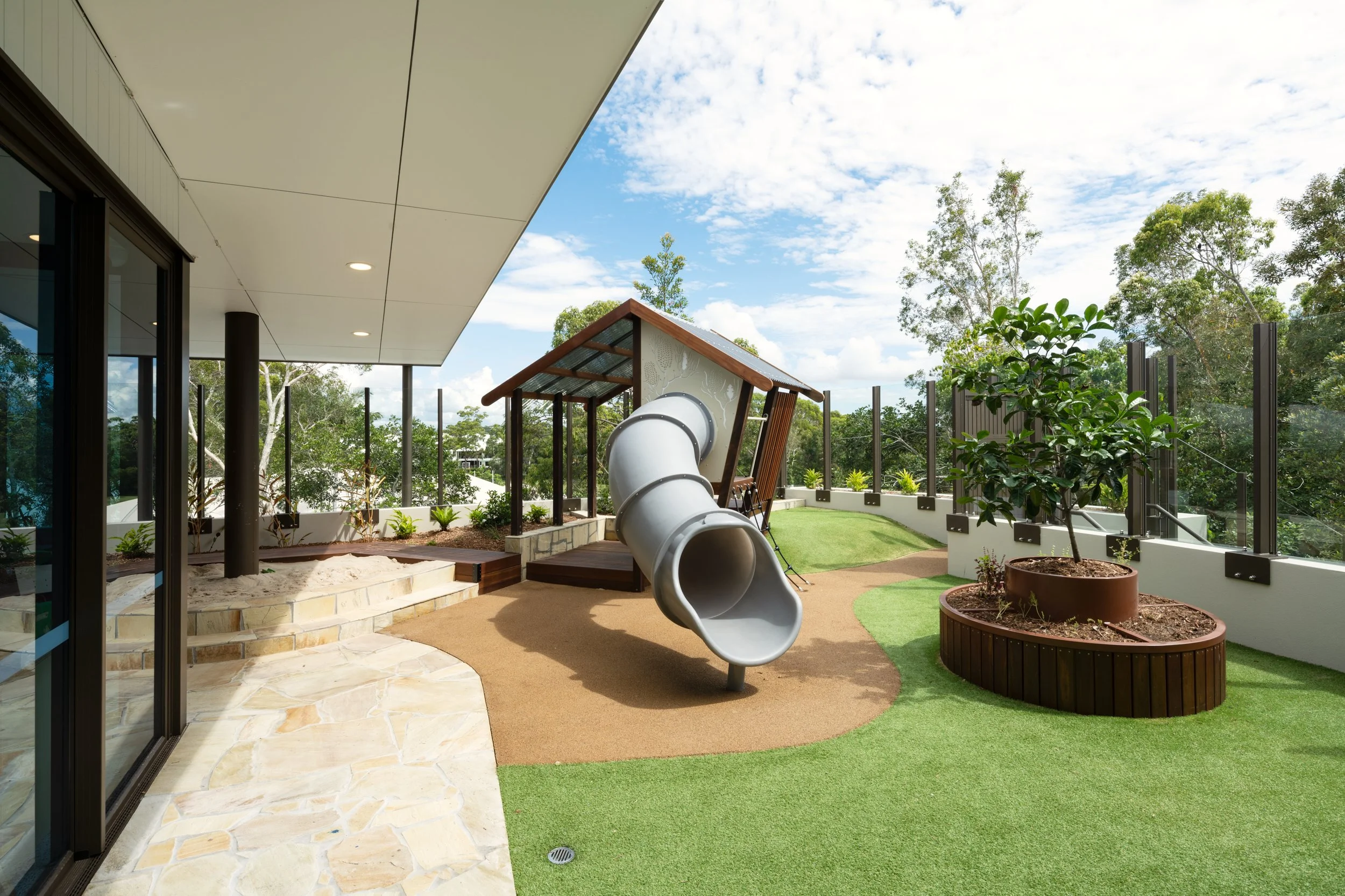 Children's outdoor play area with slide, artificial grass, potted plants, and a small house structure, on a sunny day.