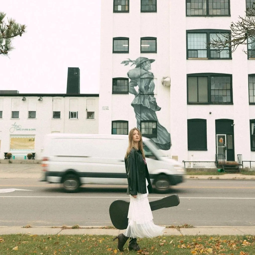 A young woman in a black leather jacket, white dress, and black boots walking on a sidewalk in front of a white building with a large mural of a woman in a hat painted on it. She is carrying a guitar case, and a white van is passing by on the street.