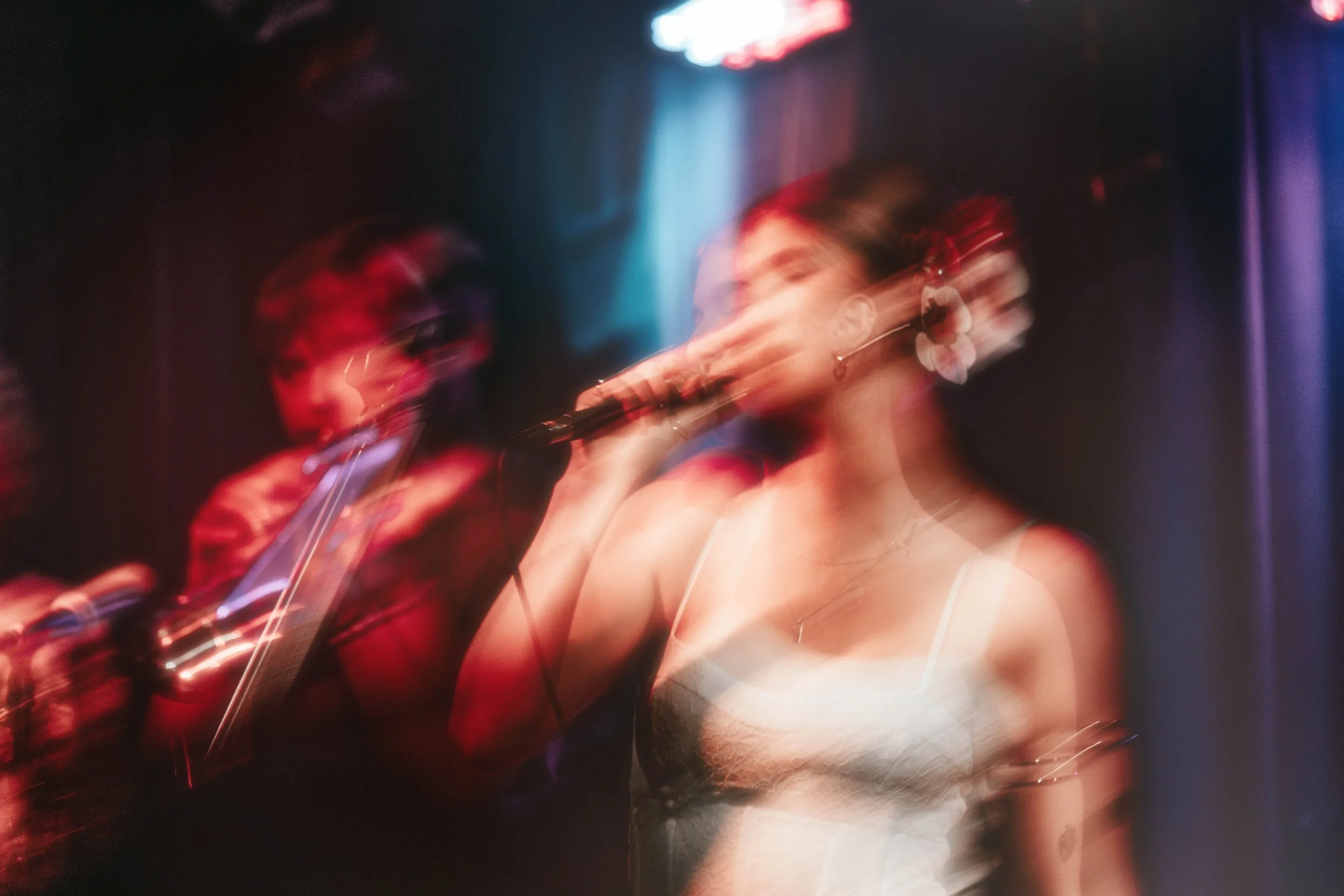 A woman singing into a microphone with a blurred background of a person playing a saxophone on a stage with colorful lights.